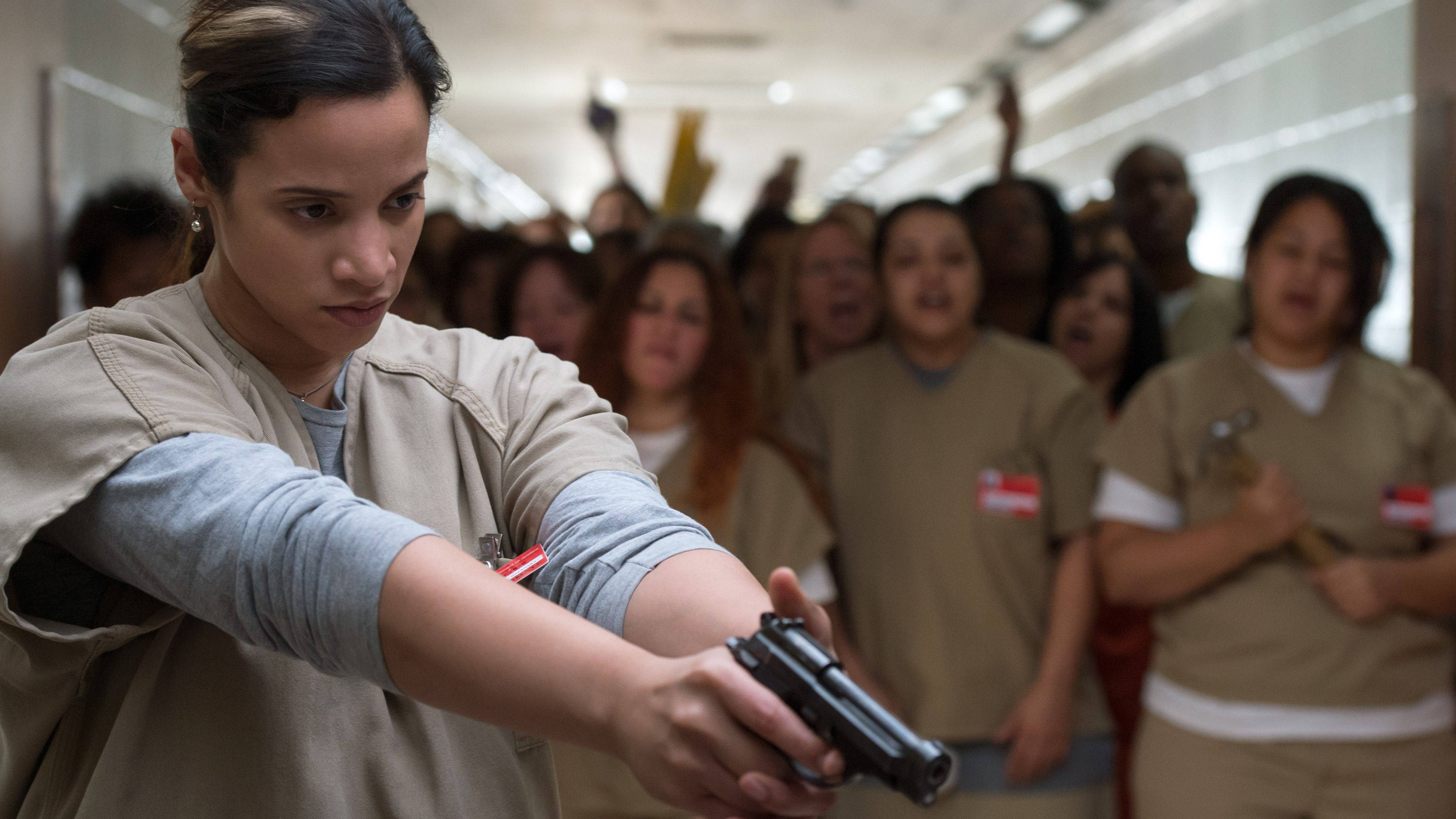 Entertainment 桌布 壁紙 — 一名身穿米色監獄制服的女人在日光燈照亮的走廊上舉起手槍瞄準，其他囚犯在她身後專注地觀看。