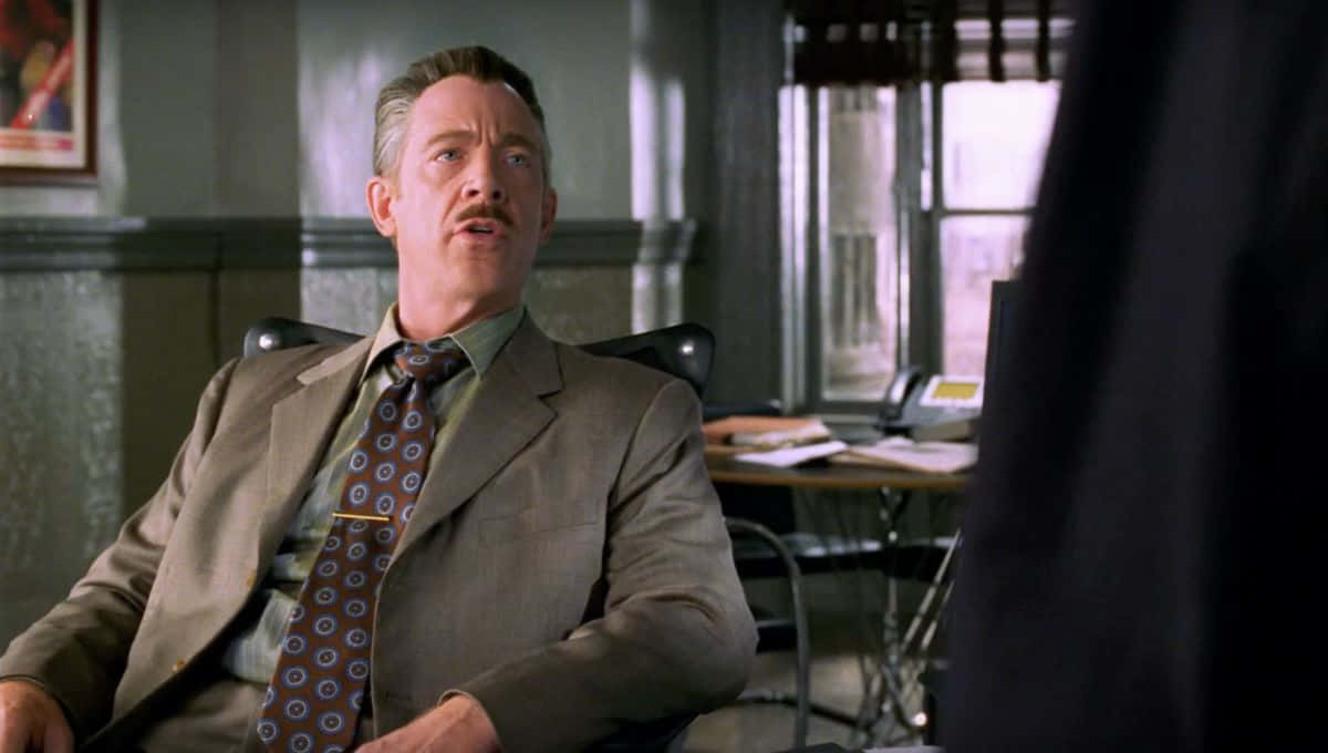 J Jonah Jameson 桌布 壁紙 — 一名身穿米色西裝和圖案領帶的男子坐在辦公椅上，表情驚訝或震驚。