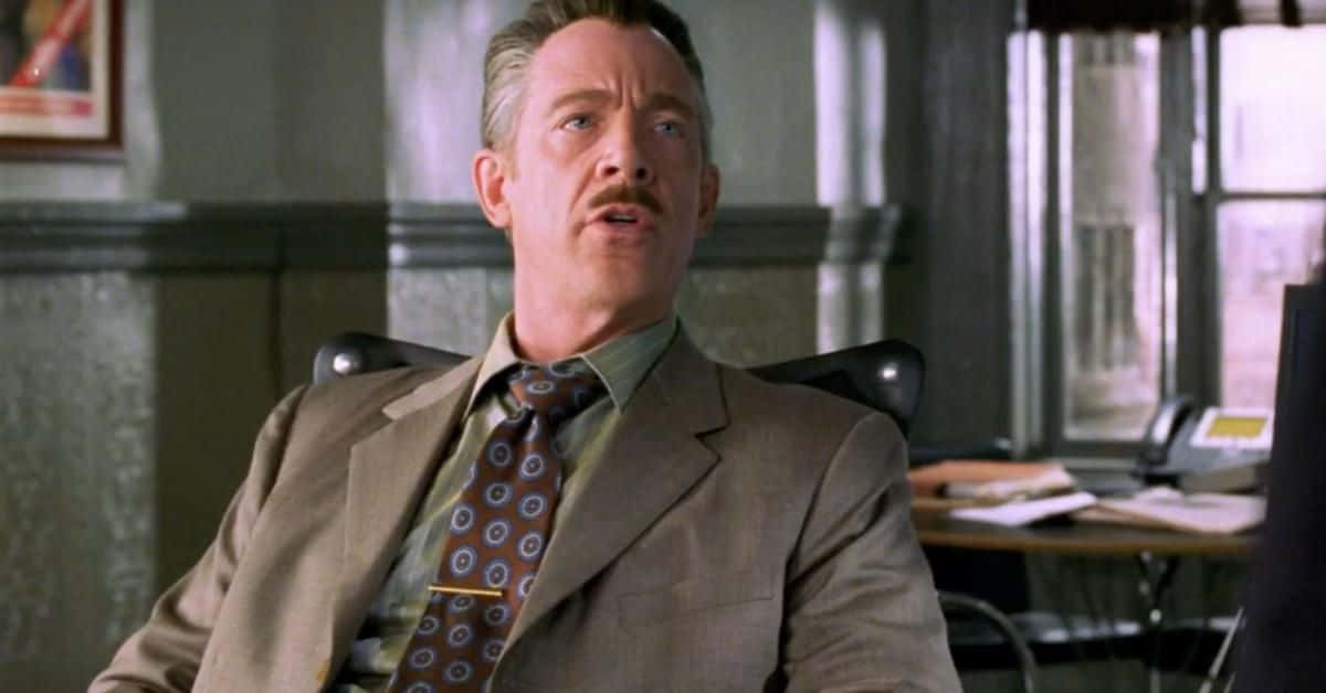 J Jonah Jameson 桌布 壁紙 — 一名身穿米色西裝和圖案領帶的男子坐在辦公椅上，表情驚訝地向上看。