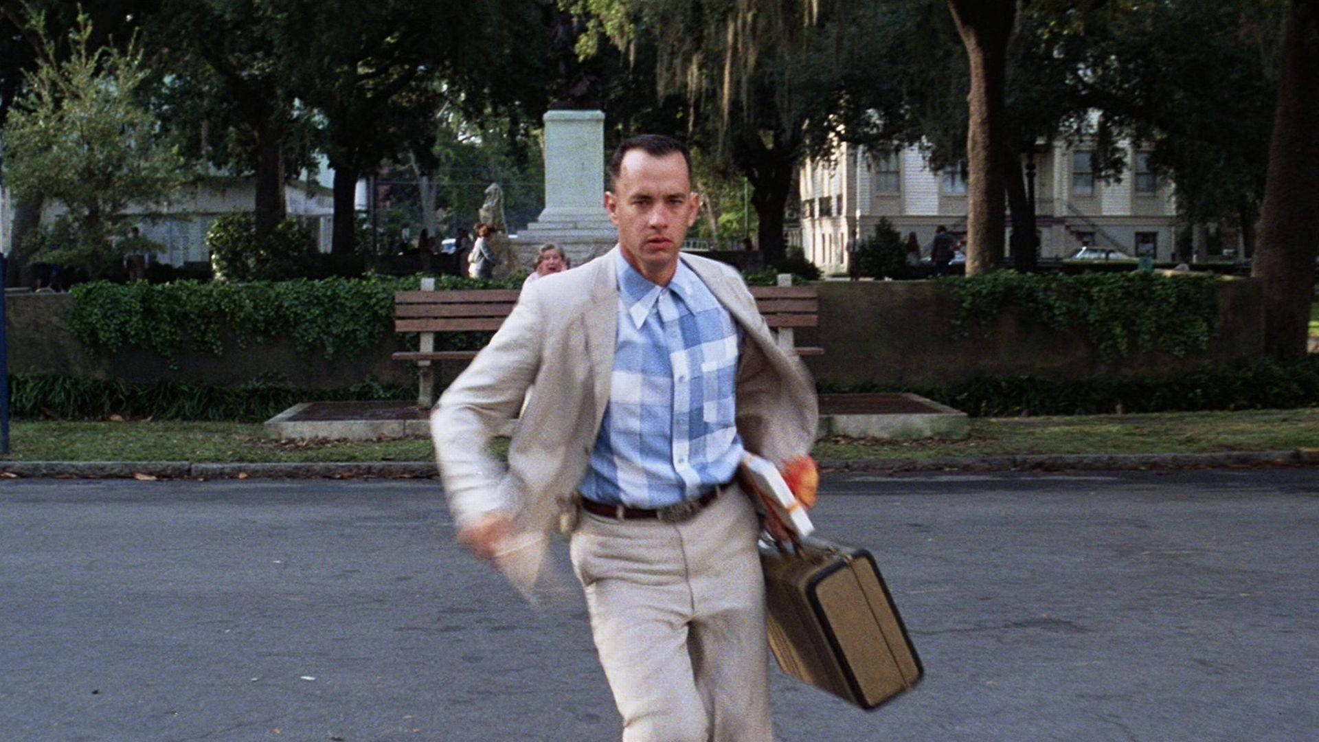 forrest 桌布 壁紙 — 一名身穿米色西裝和藍色格紋襯衫的男子，提著手提箱和文件，在綠樹成蔭的街道上匆忙奔跑。
