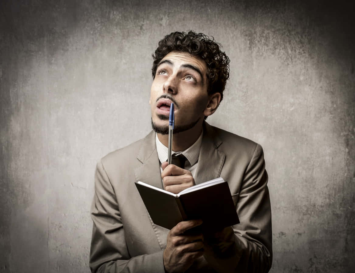 Thinking 桌布 壁紙 — 一名身穿米色西裝的男子，將筆記本和筆貼在唇邊，對著有紋理的灰色牆壁，抬頭深思。