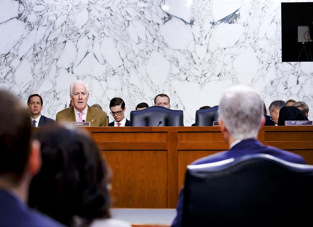 Speaker 桌布 壁紙 — 一名身穿米色西裝的男子在大理石牆前的木製聽證桌上發言，其他人從後面聆聽。