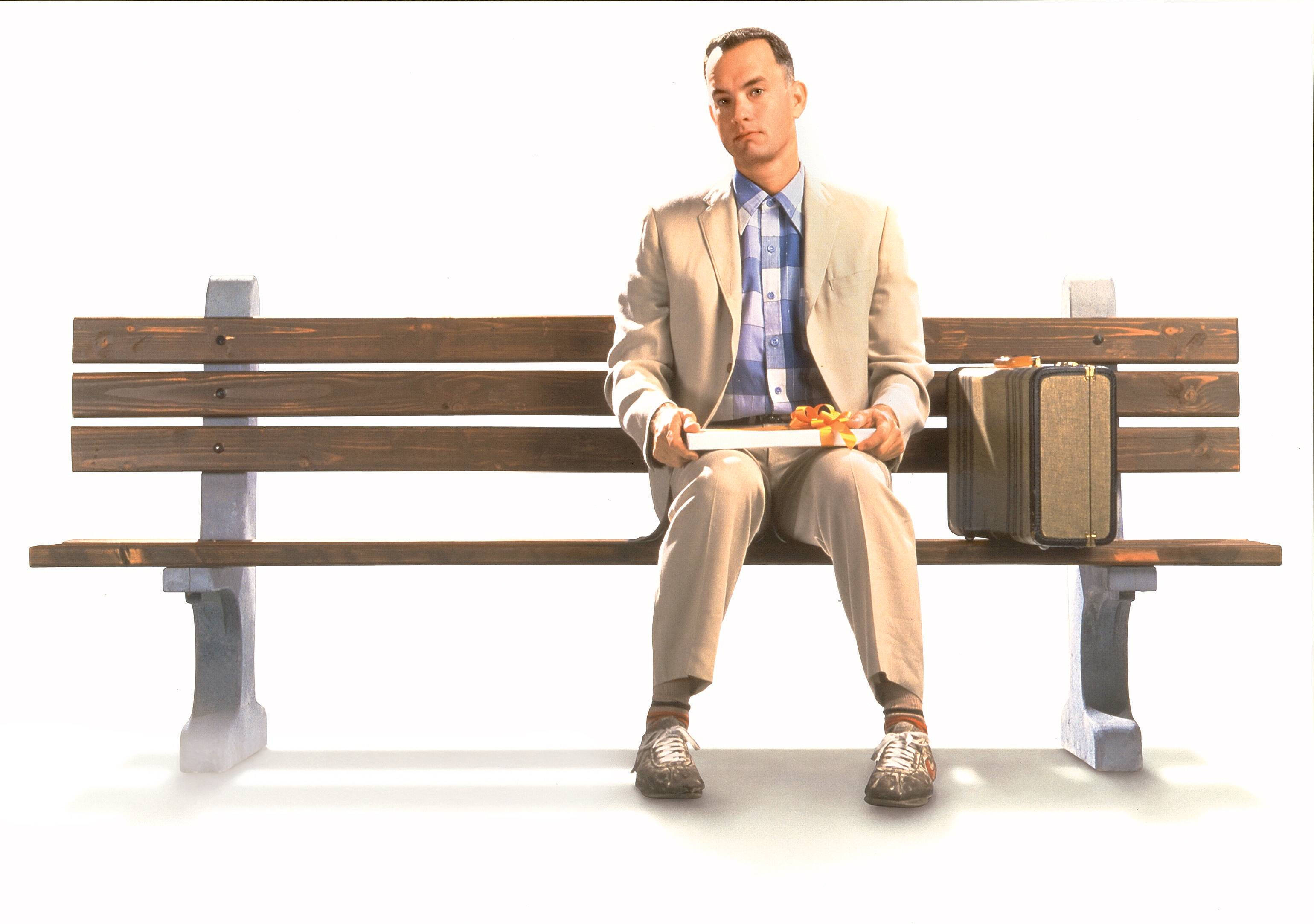 forrest 桌布 壁紙 — 一名身穿米色西裝的男子莊嚴地坐在木長椅上，手持一盤食物，旁邊放著一個復古手提箱，背景是純白色。