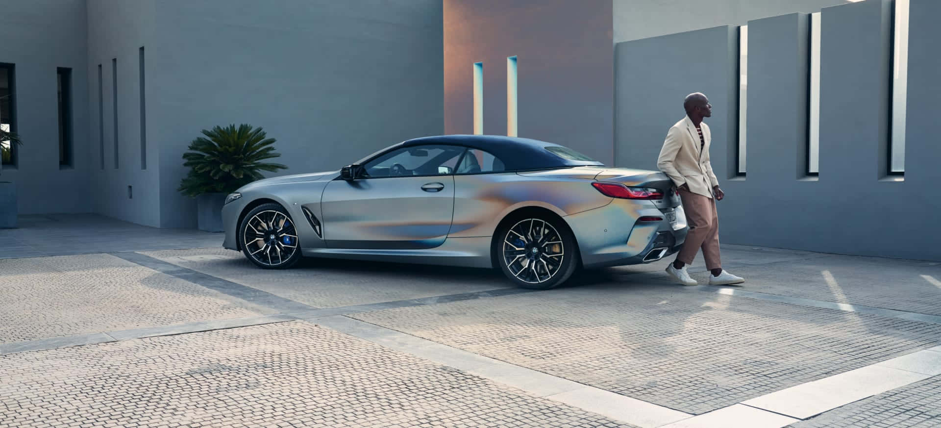 BMW 動圖 桌布 壁紙 — 一名身穿米色西裝的男子走過一輛停在現代建築風格圖案庭院中的時尚銀色敞篷車。