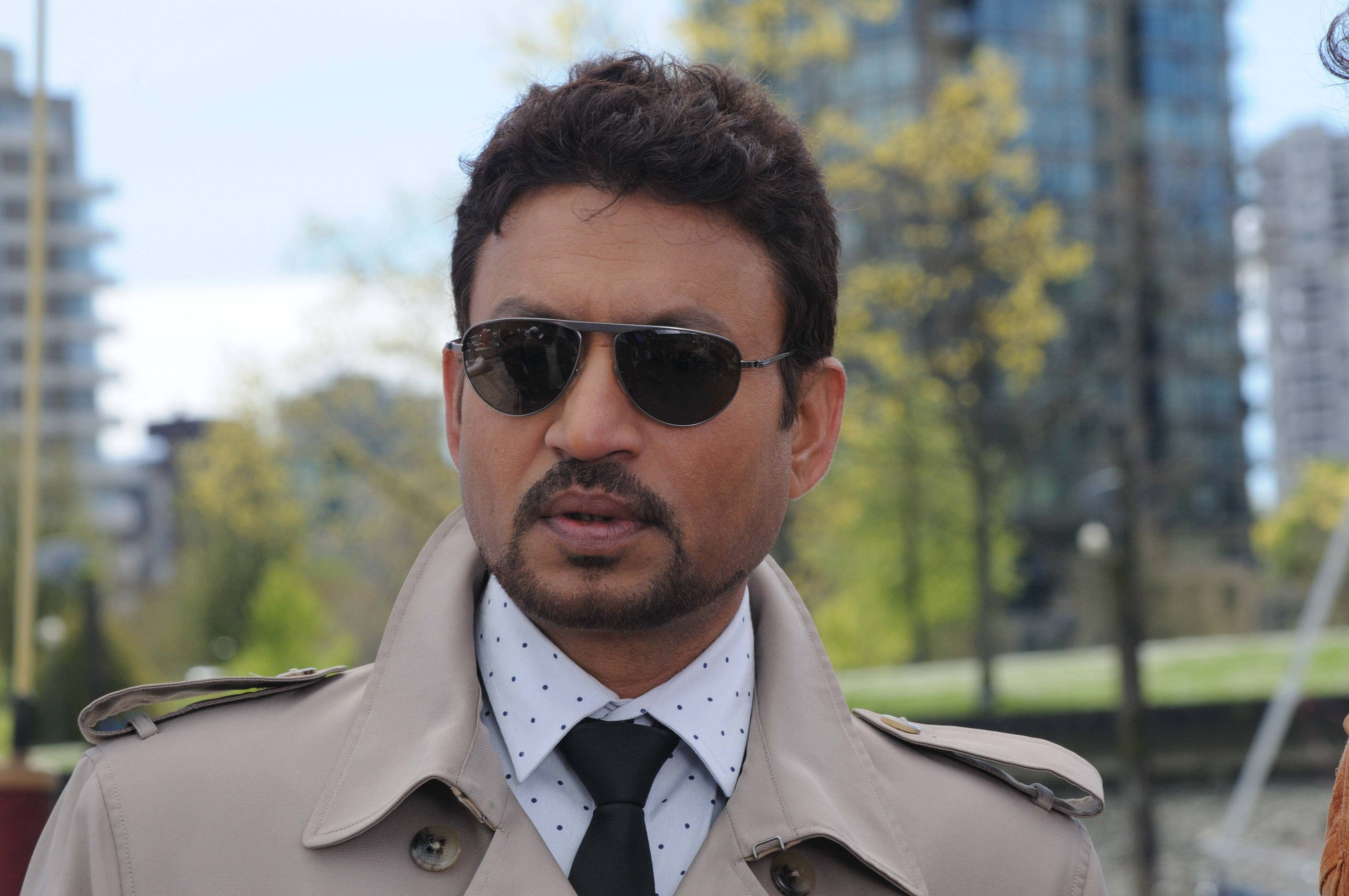 Irrfan Khan 桌布 壁紙 — 一名身穿米色風衣和太陽眼鏡的男子自信地站在模糊的城市背景下，背景有綠樹和玻璃建築。