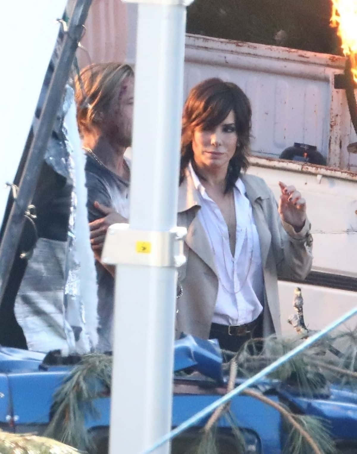 Sandra Bullock 2021 桌布 壁紙 — 一名身穿米色風衣的女子站在一名滿臉是血的男子旁邊，附近有一輛燃燒的車輛殘骸。