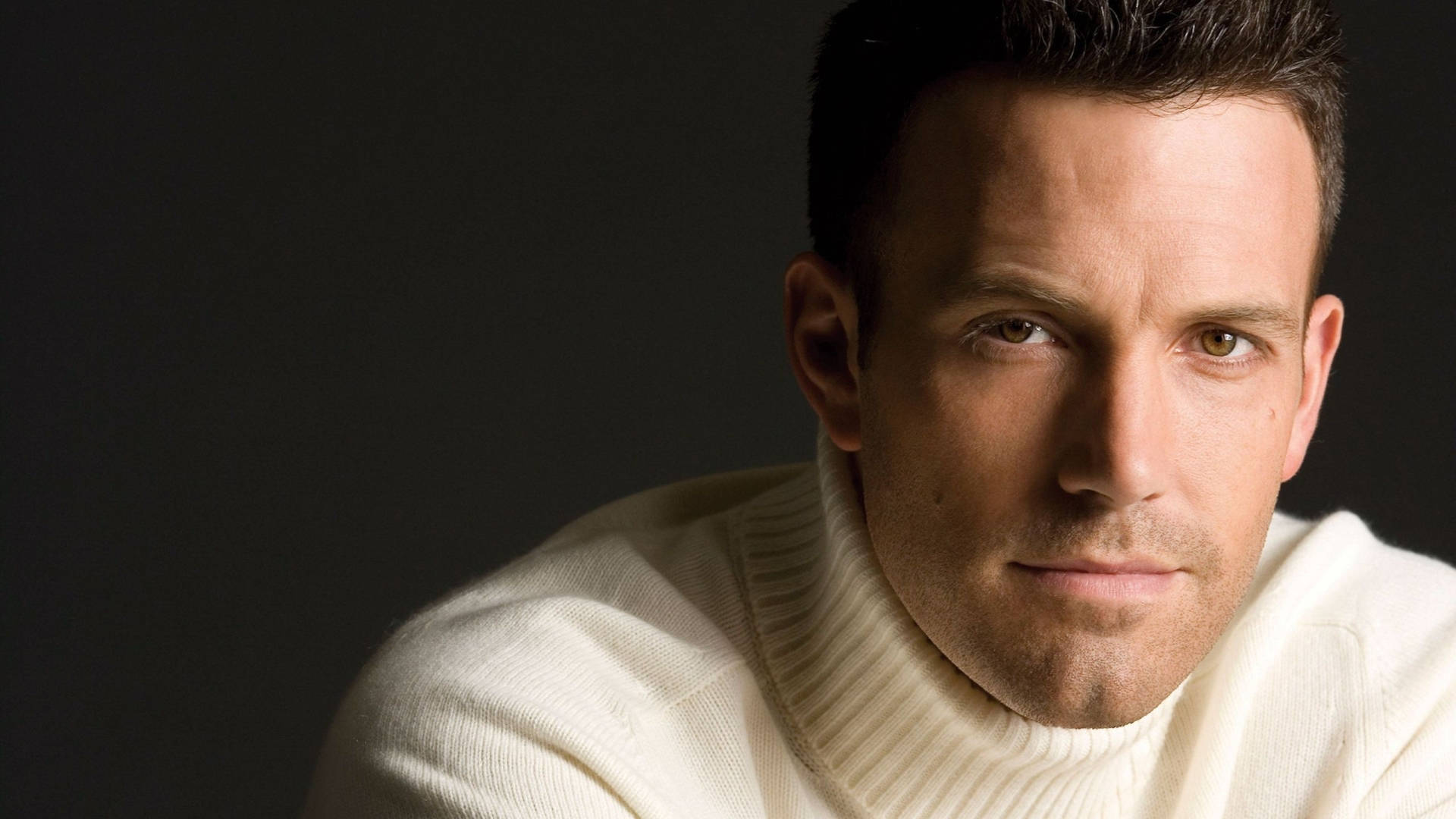 Ben Affleck 桌布 壁紙 — 一名身穿米色高領毛衣的男子在柔和的工作室燈光下，對著深色背景專注地凝視。