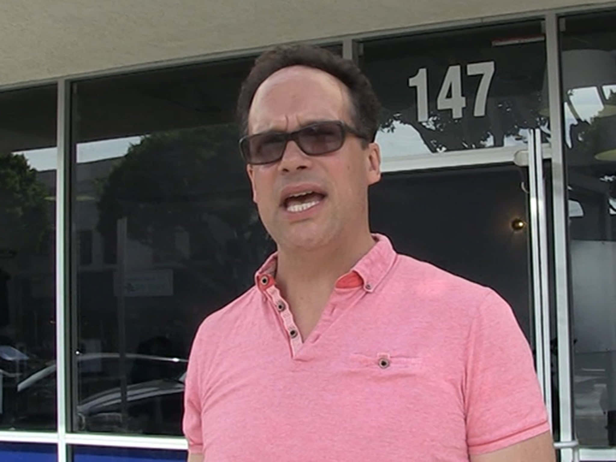Diedrich Bader 桌布 壁紙 — 一名身穿粉紅色Polo衫和太陽眼鏡的男子站在一棟玻璃門上有數字147的建築物外。