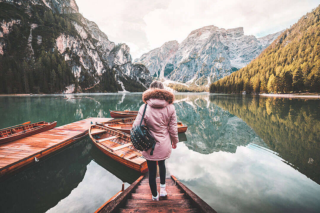 HD Scenery 桌布 壁紙 — 一名身穿粉紅色外套的人站在木碼頭上，凝視著寧靜的山湖，湖面倒映著高聳的山峰和森林覆蓋的山坡。
