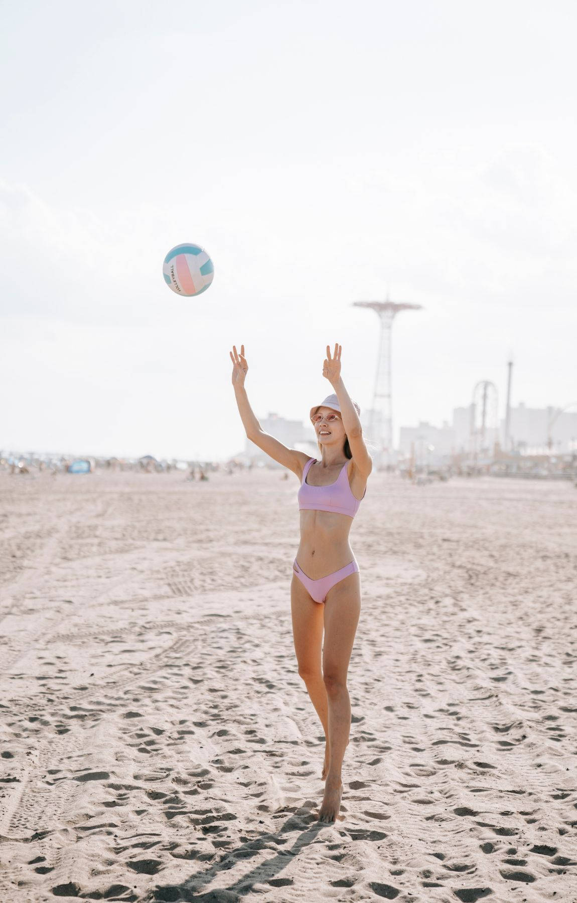 Volleyball Aesthetic 桌布 壁紙 — 一名身穿粉紅色比基尼的女子在陽光明媚的海灘上跳起擊打排球，背景朦朧中可見碼頭和遊樂園。