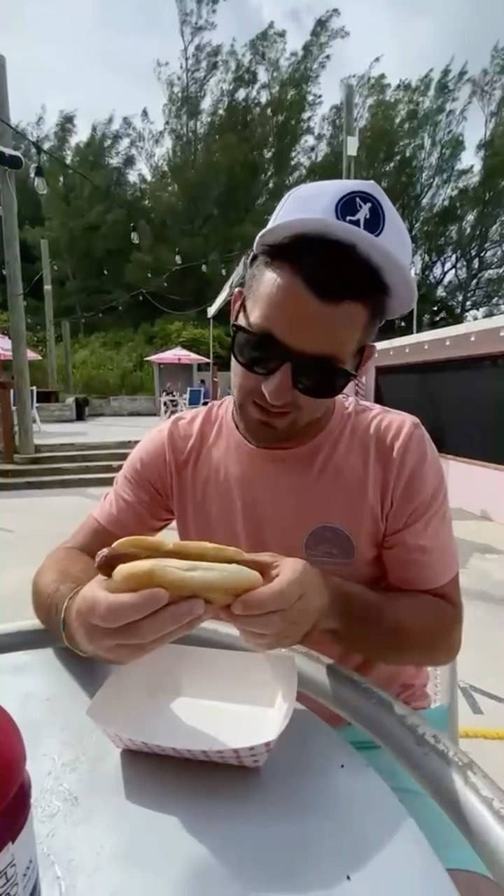 Hot Dog 桌布 壁紙 — 一名身穿粉紅色襯衫和白色帽子的男士在明亮的日光下於戶外桌子吃熱狗。