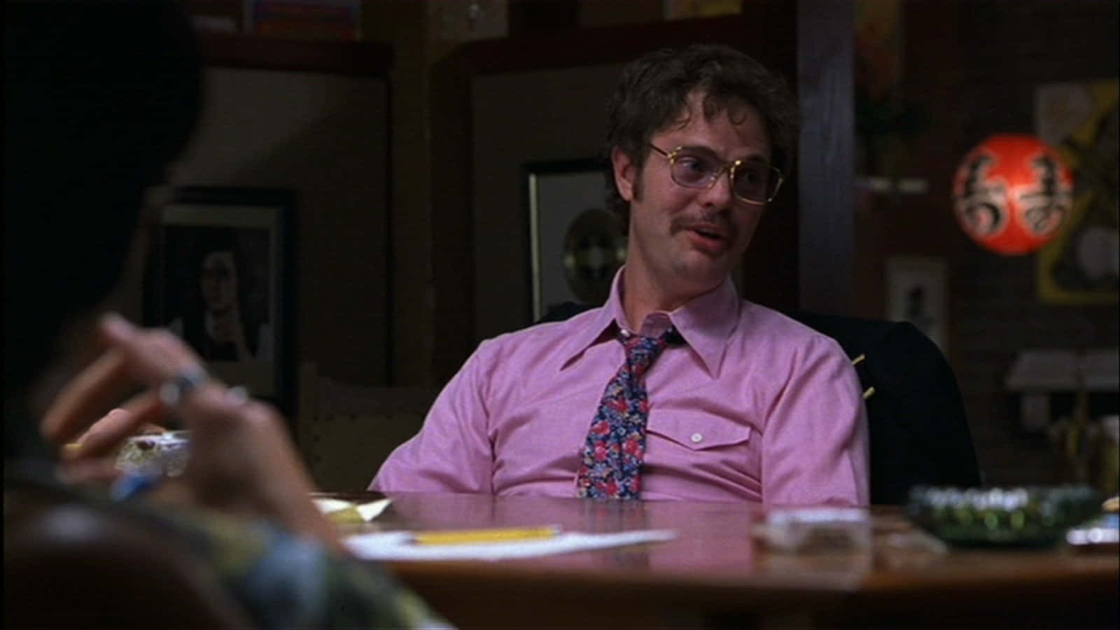 rainn wilson 桌布 壁紙 — 一名身穿粉紅色襯衫和花卉領帶的男子坐在木桌旁，房間光線昏暗，牆上掛著裱框照片和紅色燈籠。