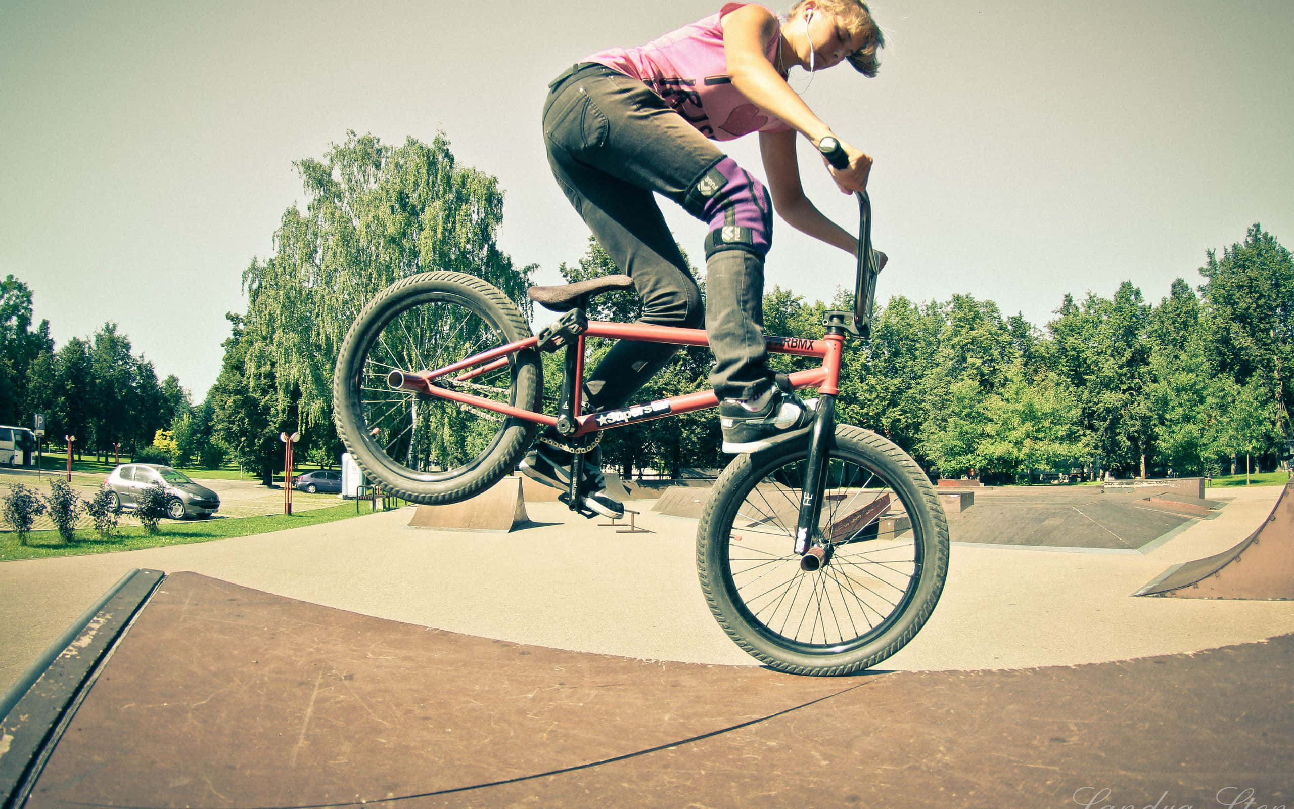 bmx 桌布 壁紙 — 一名身穿粉紅色襯衫的BMX車手在滑板公園的坡道上空，騎著紅色腳踏車表演技巧，背景有綠樹。