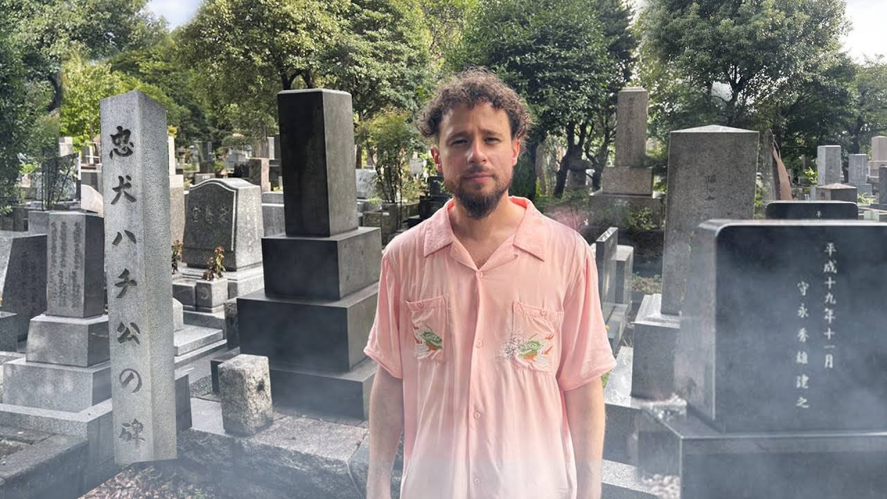 日本 桌布 壁紙 — 一名身穿粉紅色襯衫的男子，在綠樹成蔭的日本墓地中，莊嚴地站在石墓碑之間。