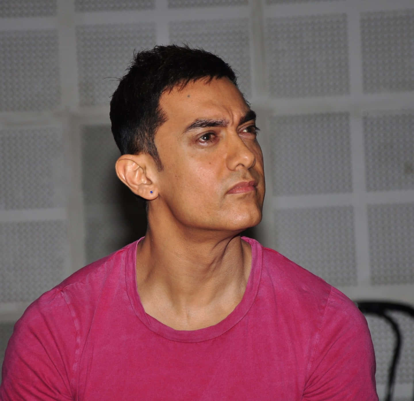 Aamir Khan 桌布 壁紙 — 一名身穿粉紅色襯衫的男子，戴著低調的藍色耳環，若有所思地凝視著網格圖案的牆壁。