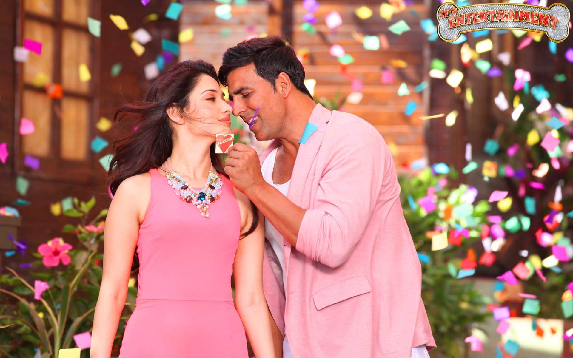 Akshay Kumar 桌布 壁紙 — 一名身穿粉紅色西裝外套的男子，頑皮地餵一名身穿粉紅色連衣裙的女子吃棒棒糖，五彩紙屑在他們周圍飄落。