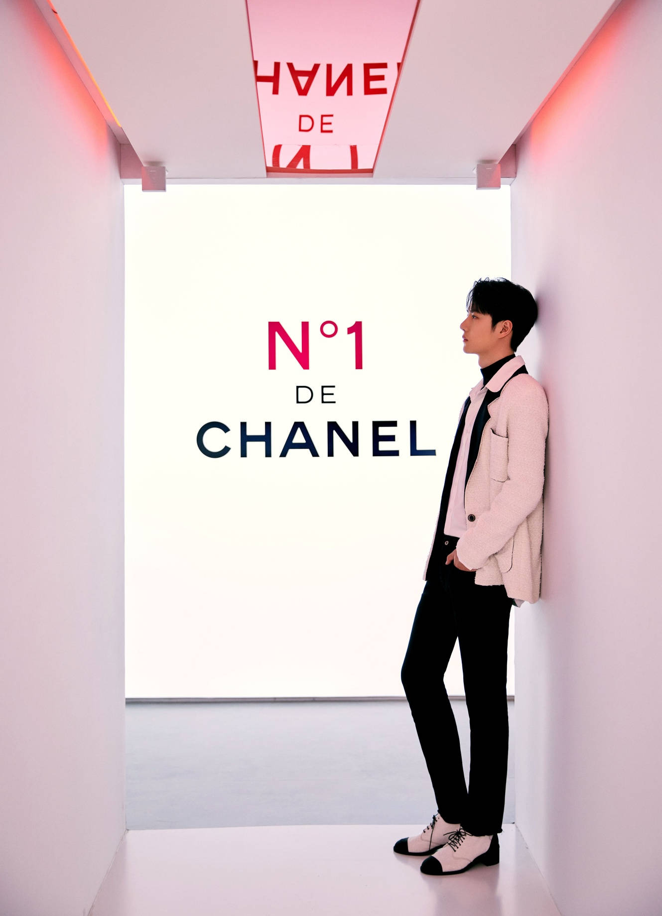 王一博 桌布 壁紙 — 一名身穿粉紅色西裝外套的男子靠在發光紅色 Chanel 標誌下的簡約白色走廊牆上。