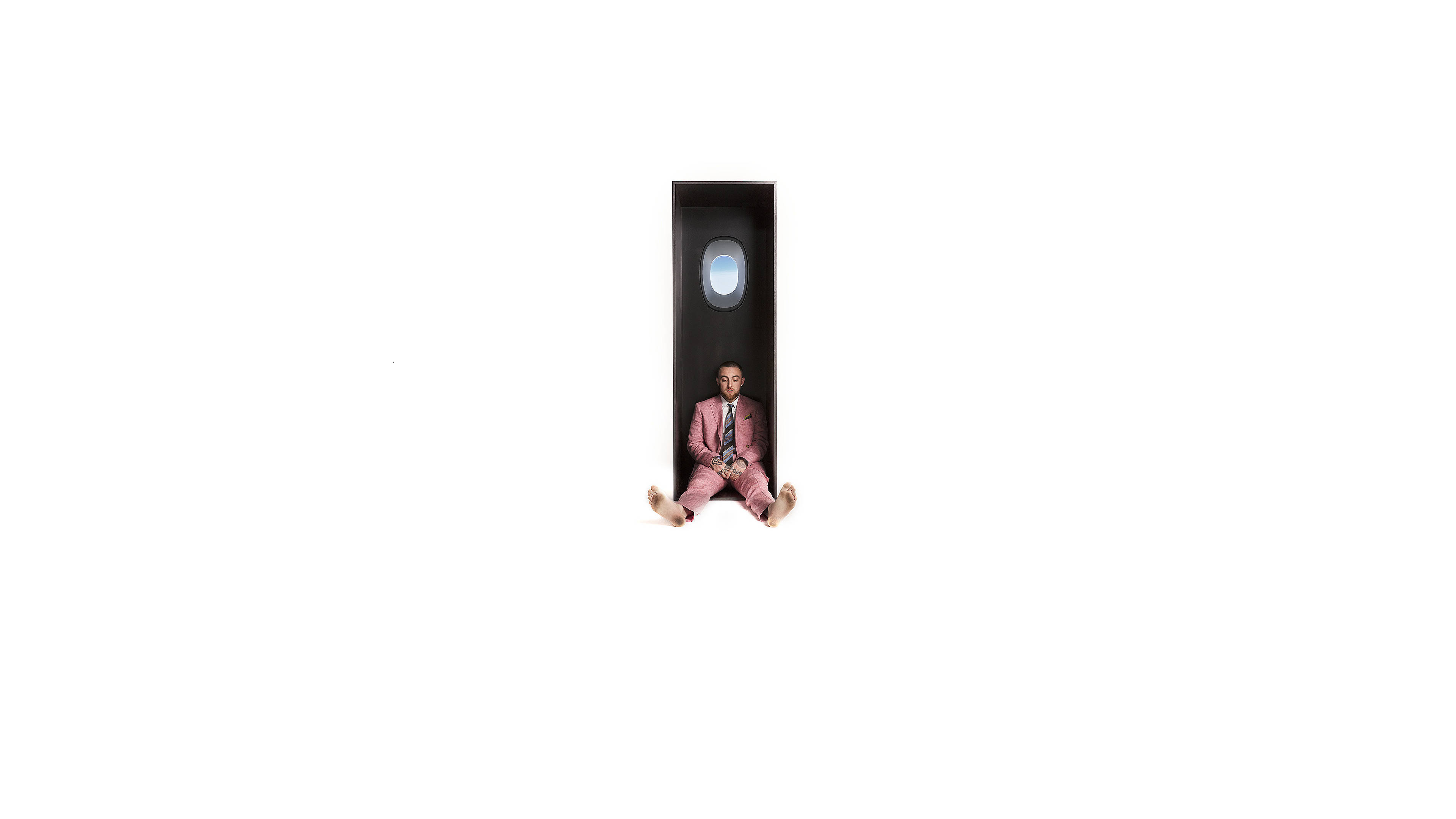 mac miller 4K 桌布 壁紙 — 一名身穿粉紅色西裝的男子赤腳坐在狹窄的飛機艙內，艙內有圓形窗戶，背景為純白色。
