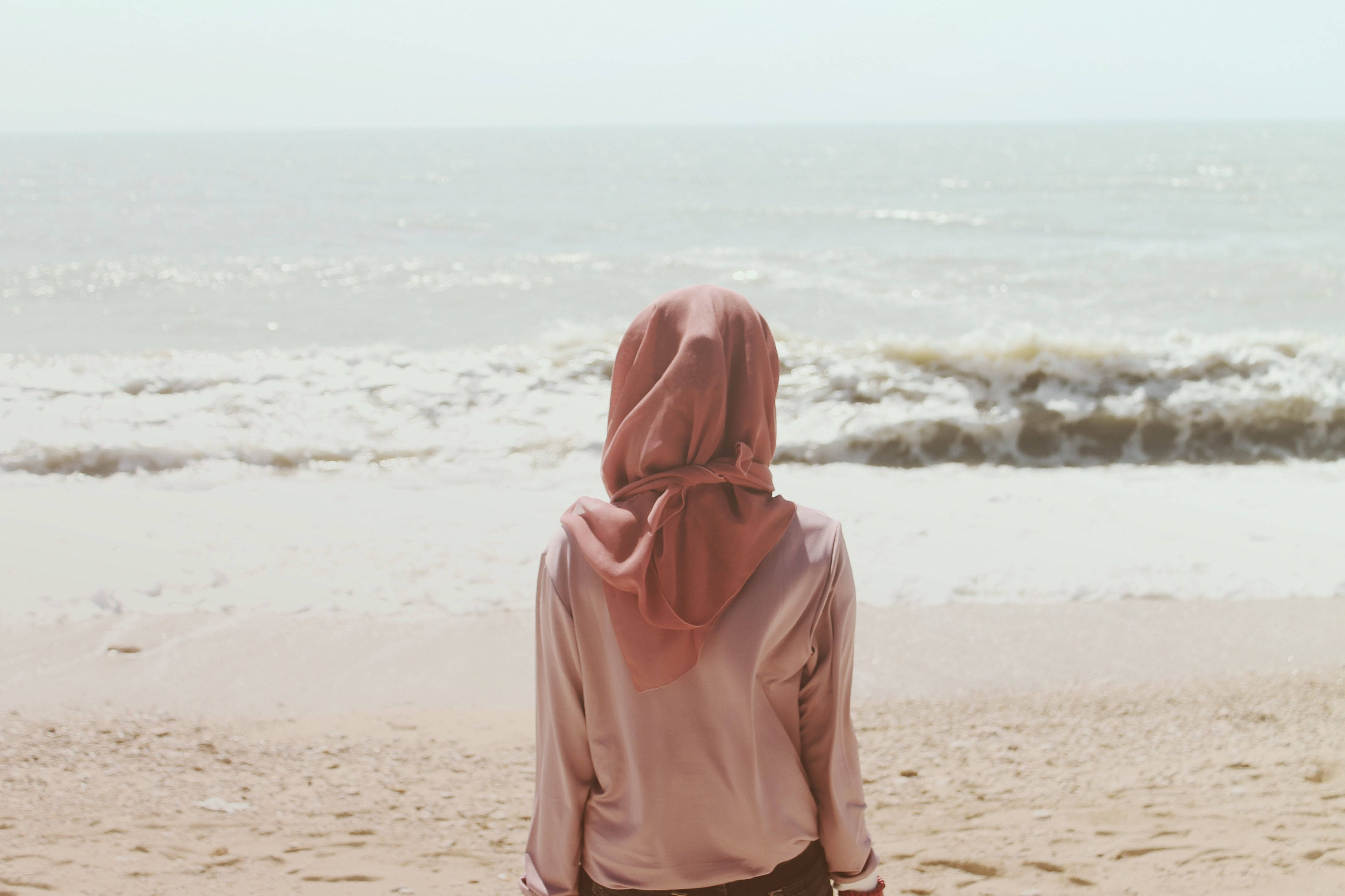 muslim 桌布 壁紙 — 一名身穿粉紅色頭巾的人獨自站在沙灘上，凝視著朦朧天空下的海浪。