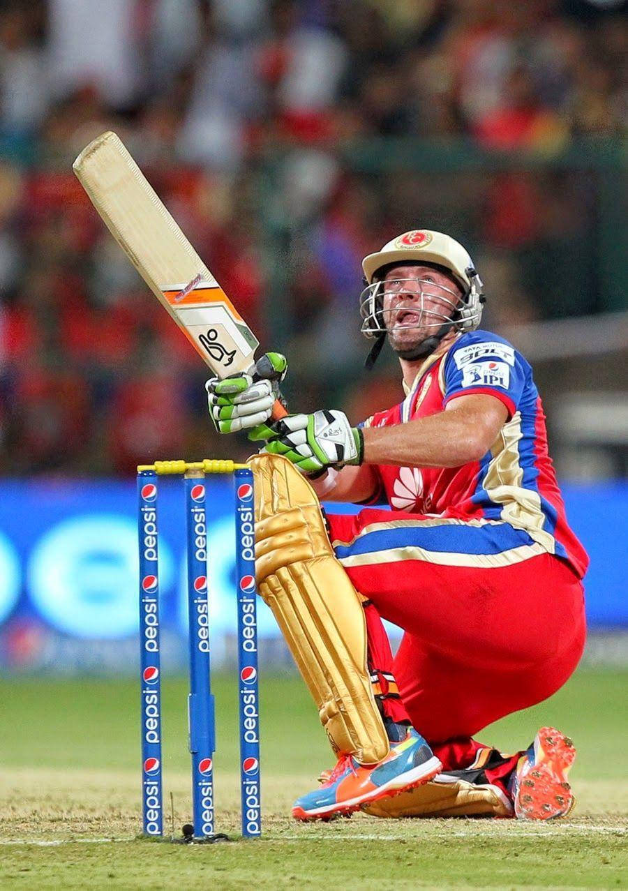Ab De Villiers 桌布 壁紙 — 一名身穿紅、藍、金色球衣的板球運動員在球場燈光下，跪在門柱旁，手持球棒，展現勝利姿態。