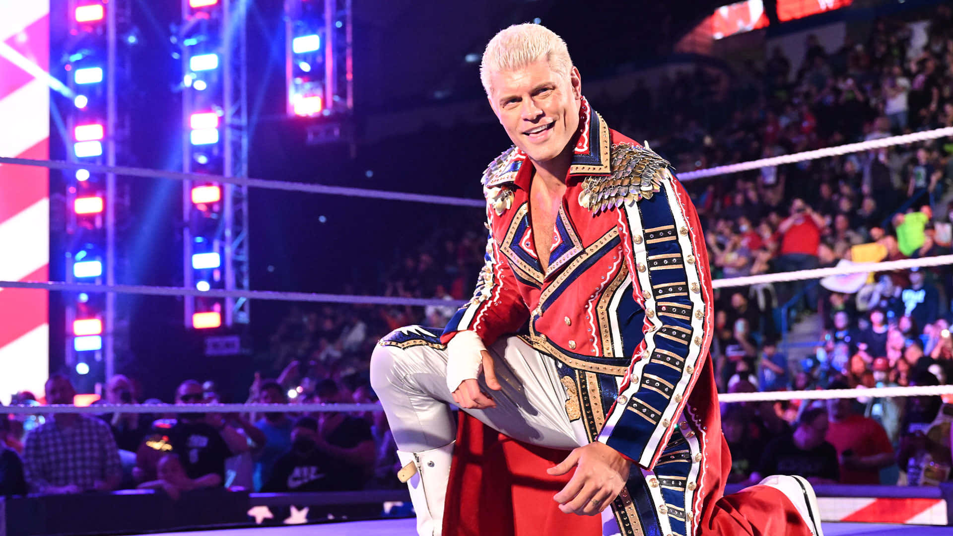 WWE 桌布 壁紙 — 一名身穿紅、金、藍色華麗服裝的摔角手，在燈光明亮的擂台上自信地跪下，身後是歡呼的人群。