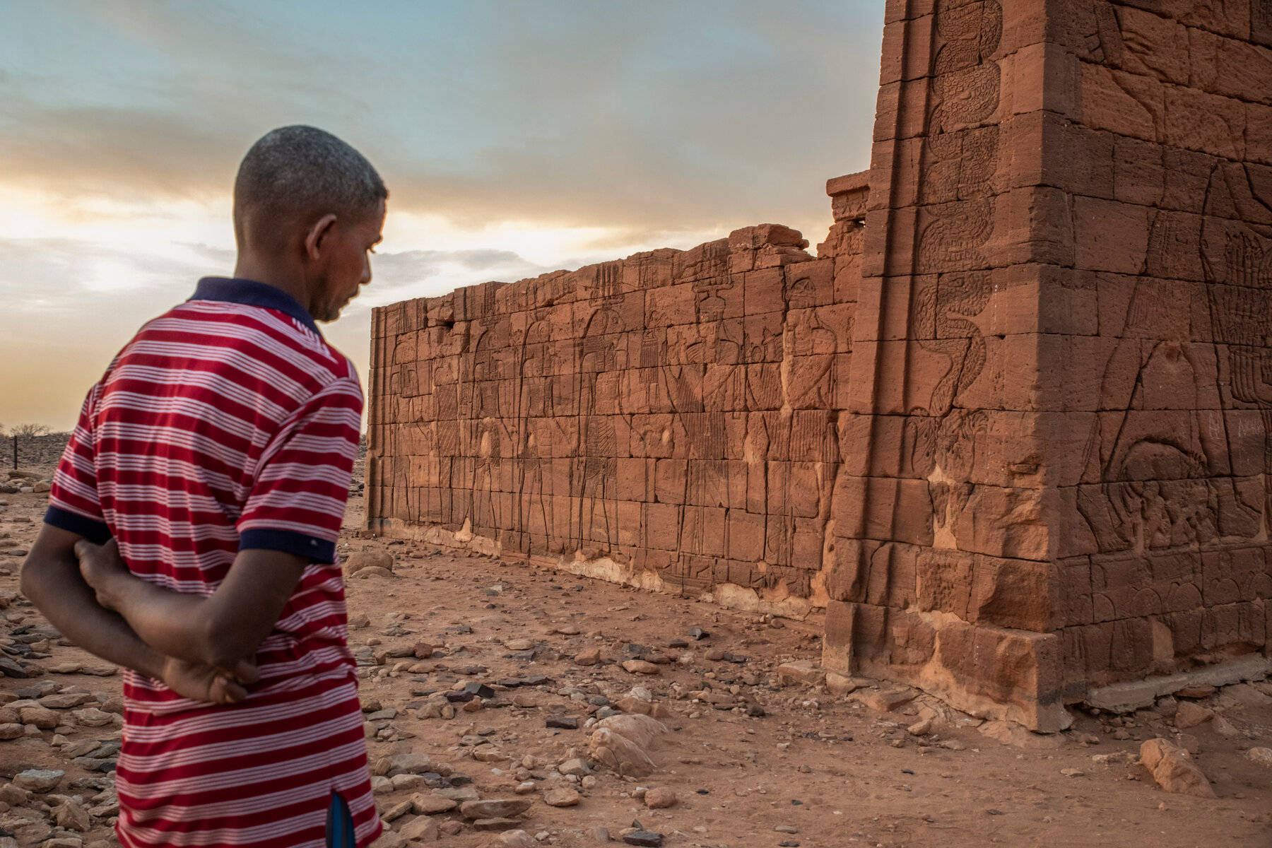 Sudan 桌布 壁紙 — 一名身穿紅條紋襯衫的男子站在古老的雕刻石頭廢墟前，背景是柔和的金色日落天空。