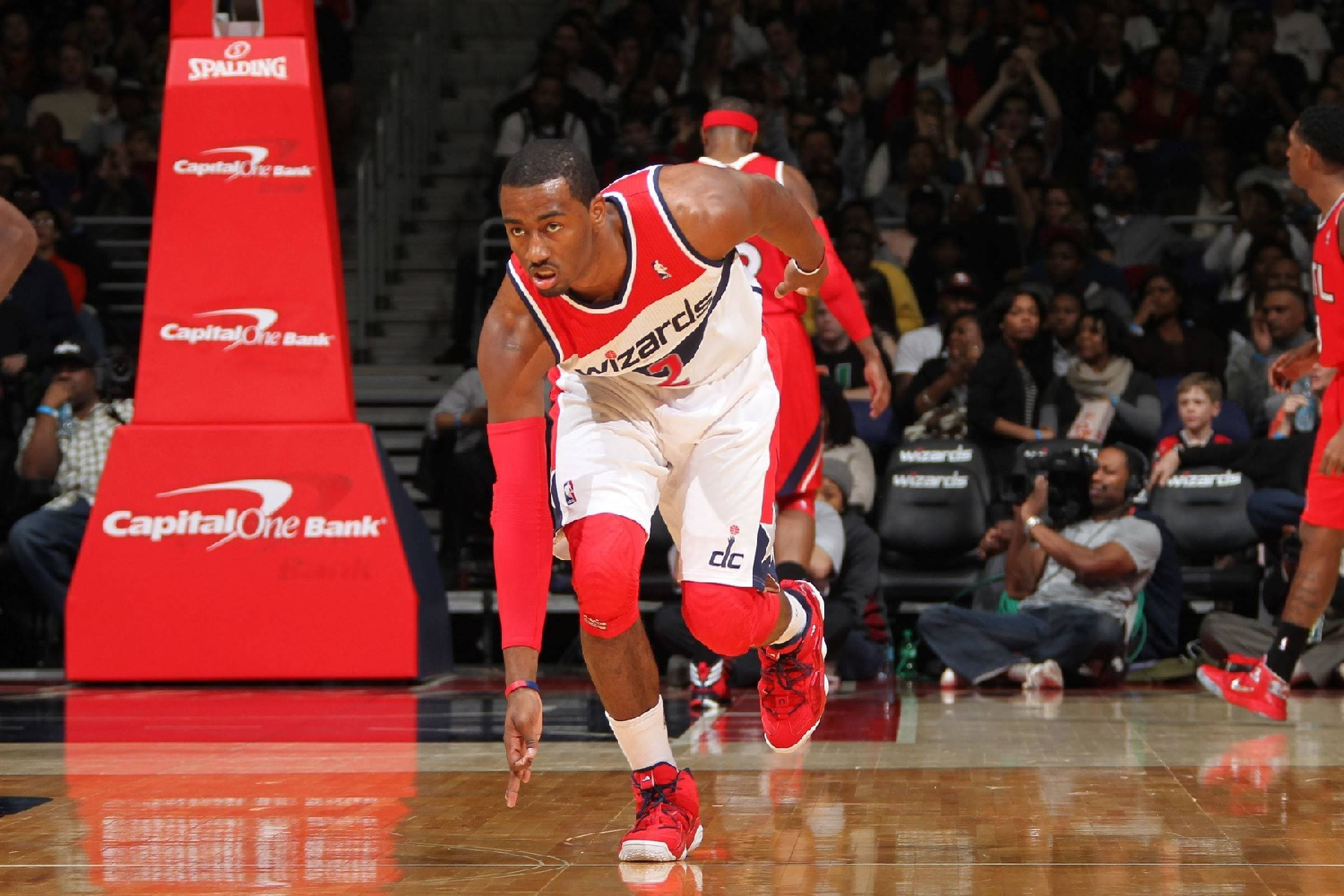 John Wall 桌布 壁紙 — 一名身穿紅白巫師隊球衣的籃球運動員在拋光場地上衝刺，觀眾和Capital One品牌標誌清晰可見。