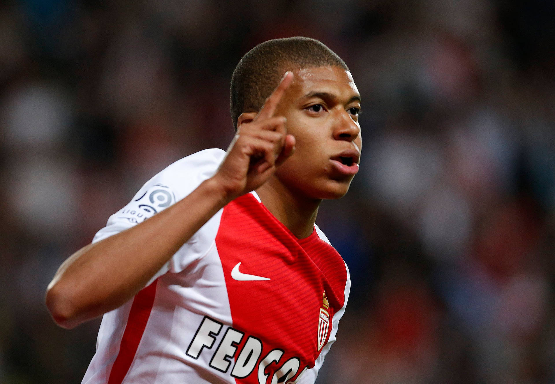 Mbappe 桌布 壁紙 — 一名身穿紅白球衣的年輕足球員在模糊的體育場人群中舉起手指慶祝。