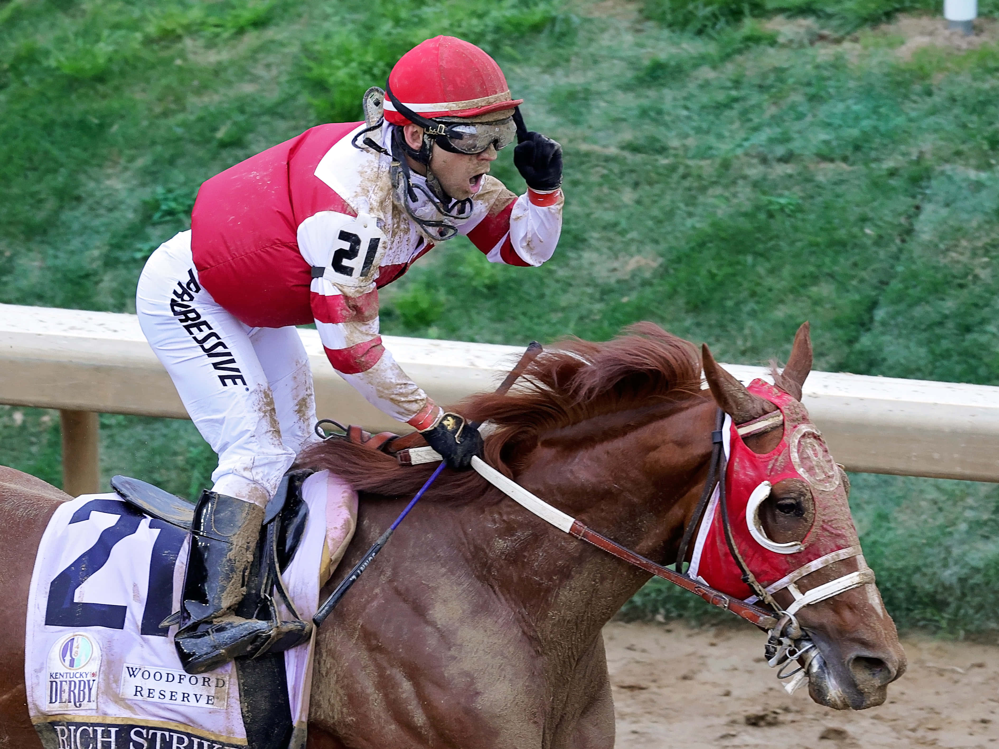 Kentucky Derby 桌布 壁紙 — 一名身穿紅白綢緞的騎師在泥地賽道上俯身向前，騎在棕色賽馬上勝利地呼喊。
