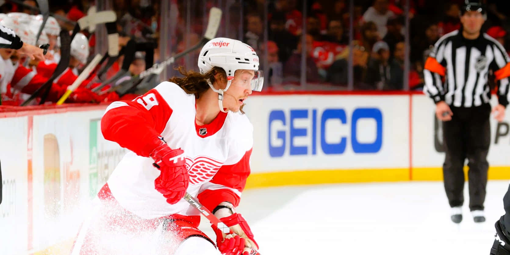 Tyler Bertuzzi 桌布 壁紙 — 一名身穿紅白裝備的冰球運動員在冰上激烈滑行，背景有裁判和人群。