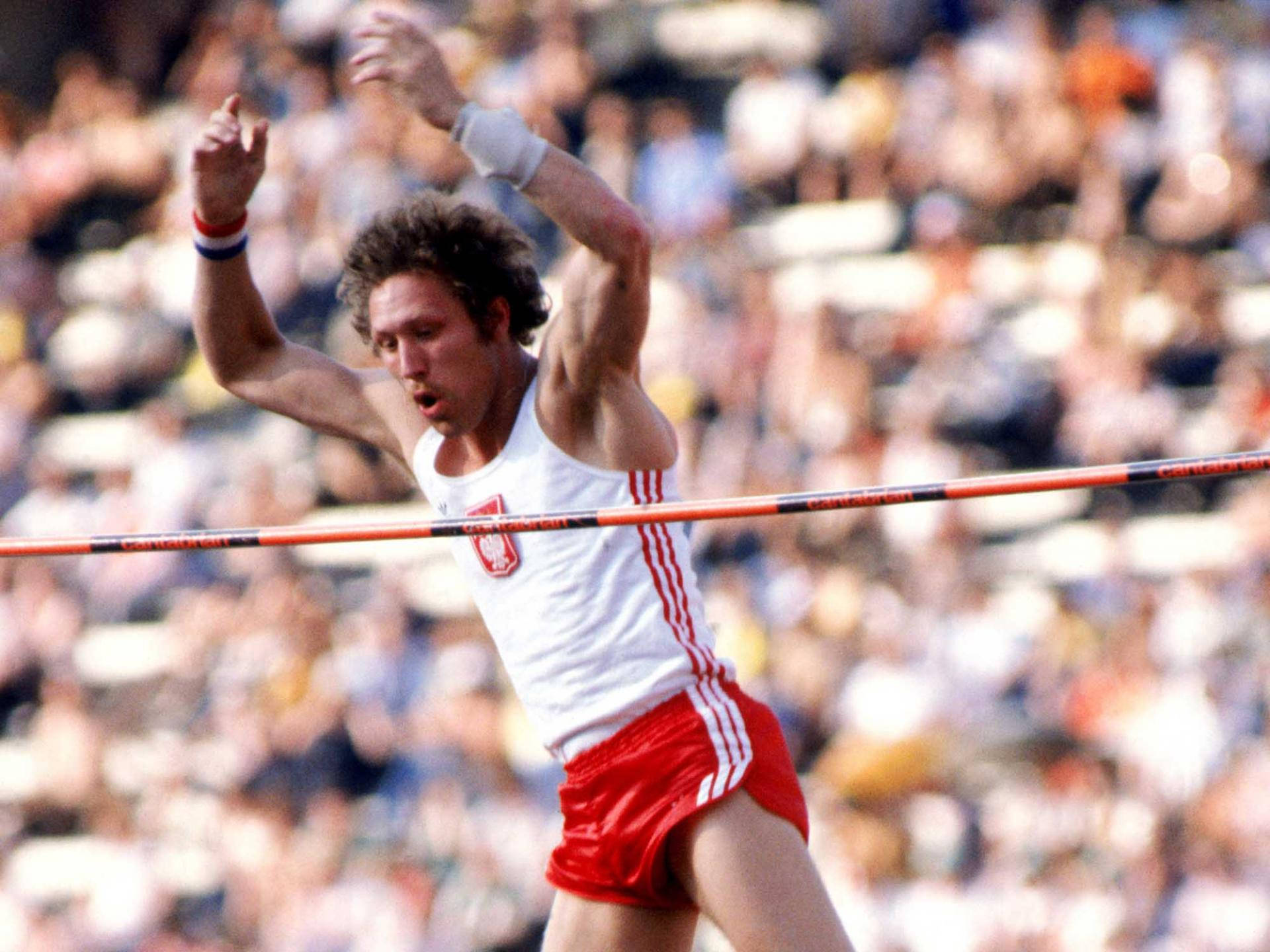 Athletic 桌布 壁紙 — 一名身穿紅白裝備的男子撐竿跳高選手，在模糊的體育場人群前，高舉雙臂越過橫桿，慶祝勝利。