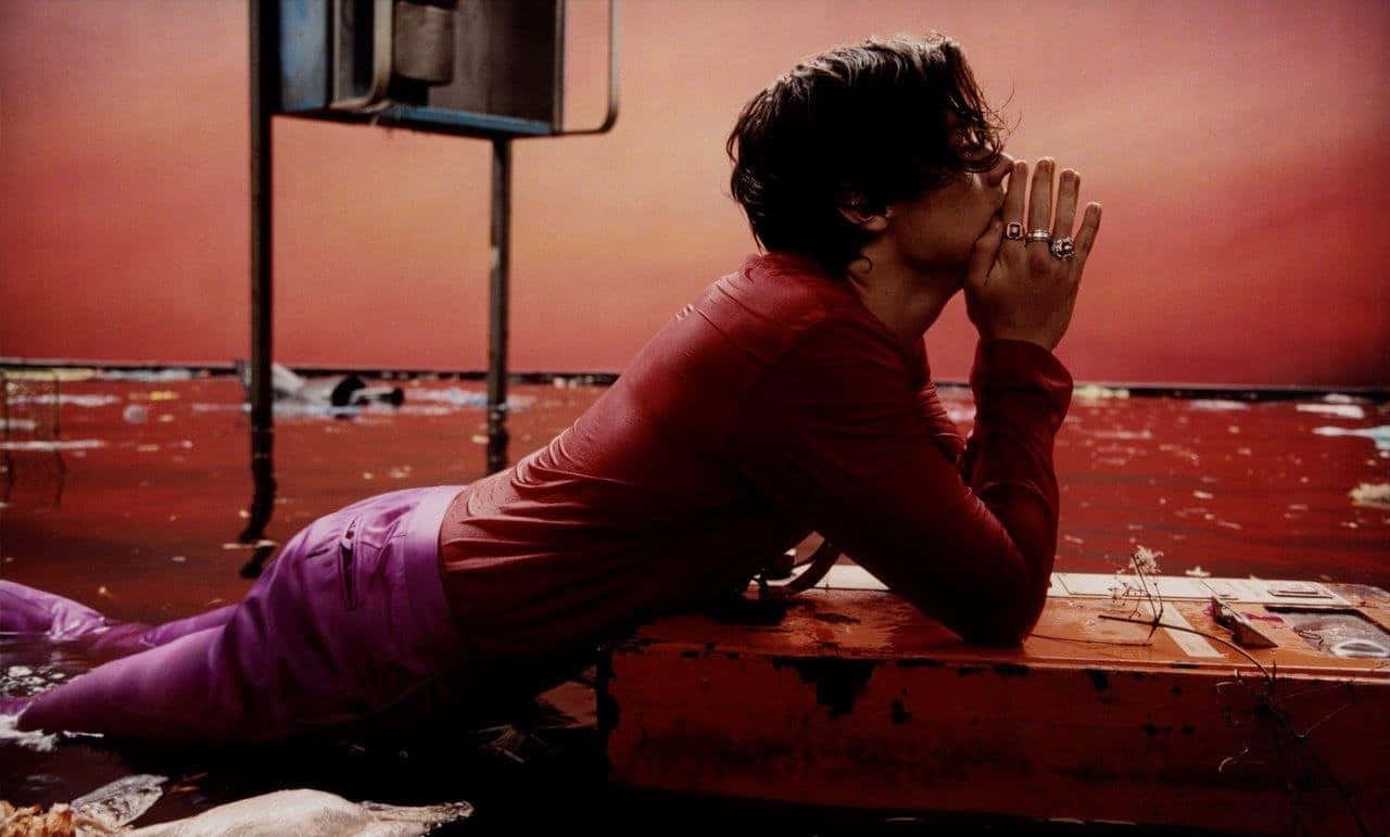 Harry Styles Laptop 桌布 壁紙 — 一名身穿紅紫衣服的人躺在一塊生鏽的金屬表面上，周圍是淹水、鏽色的城市廢墟。