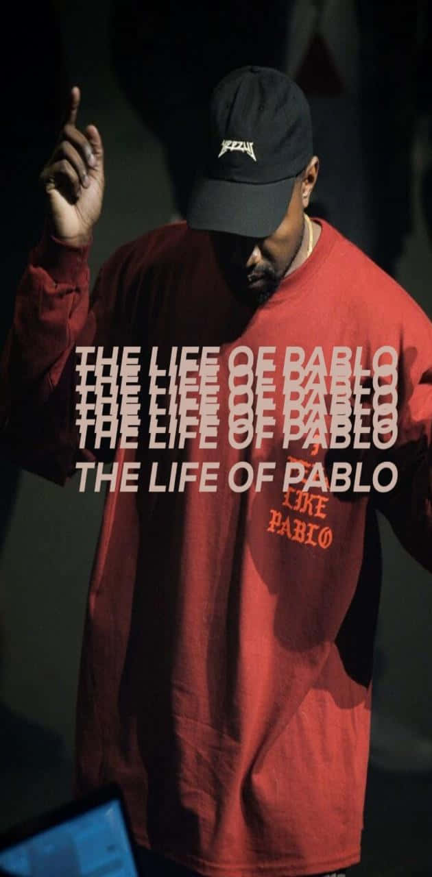 The Life Of Pablo 桌布 壁紙 — 一名身穿紅色「The Life of Pablo」衛衣和黑色鴨舌帽的男子，在戲劇性的舞台燈光下向上做手勢。