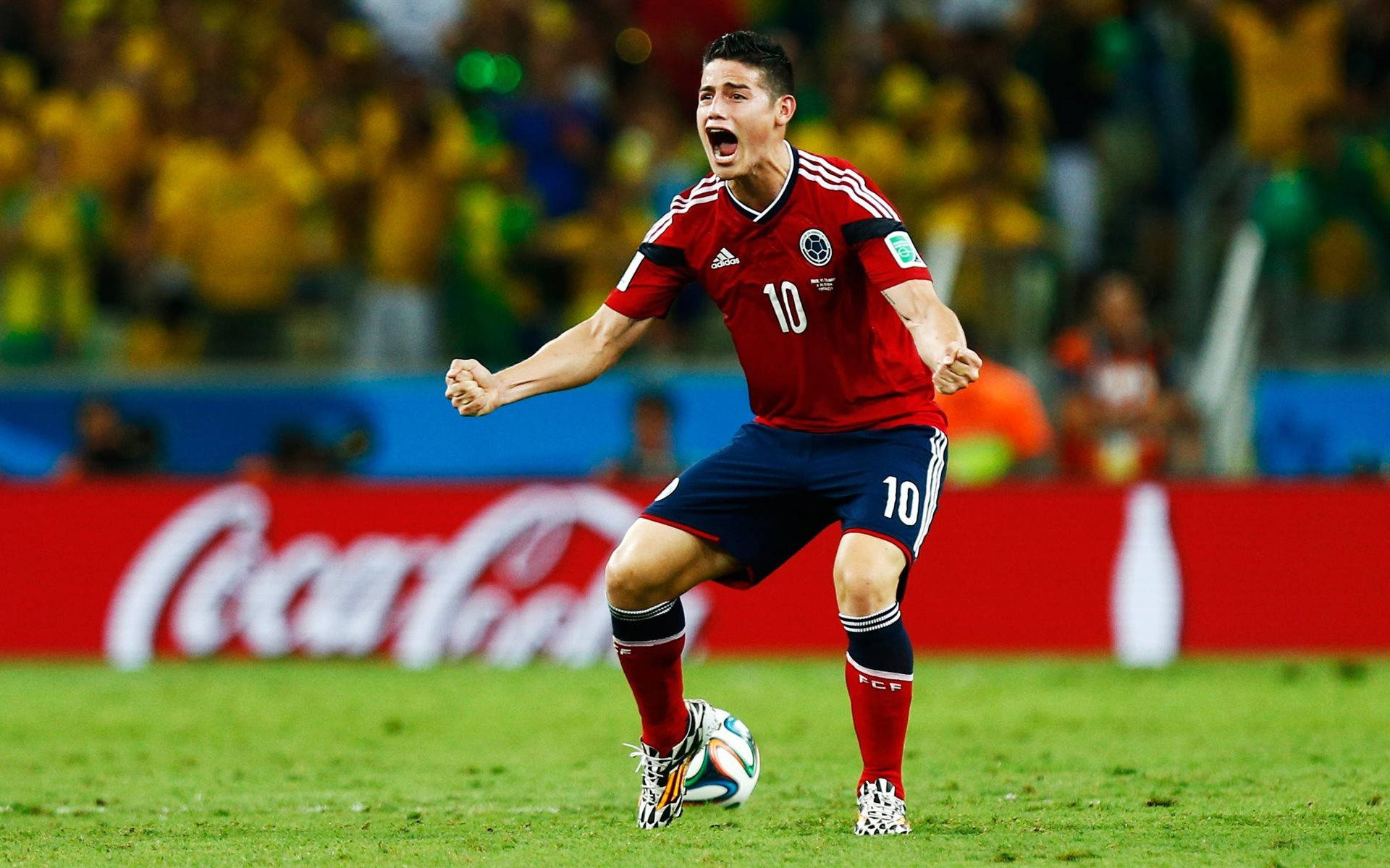 James Rodriguez 桌布 壁紙 — 一名身穿紅色10號球衣的哥倫比亞足球員在充滿活力的綠色球場上熱情慶祝，身後有黃色球迷歡呼。