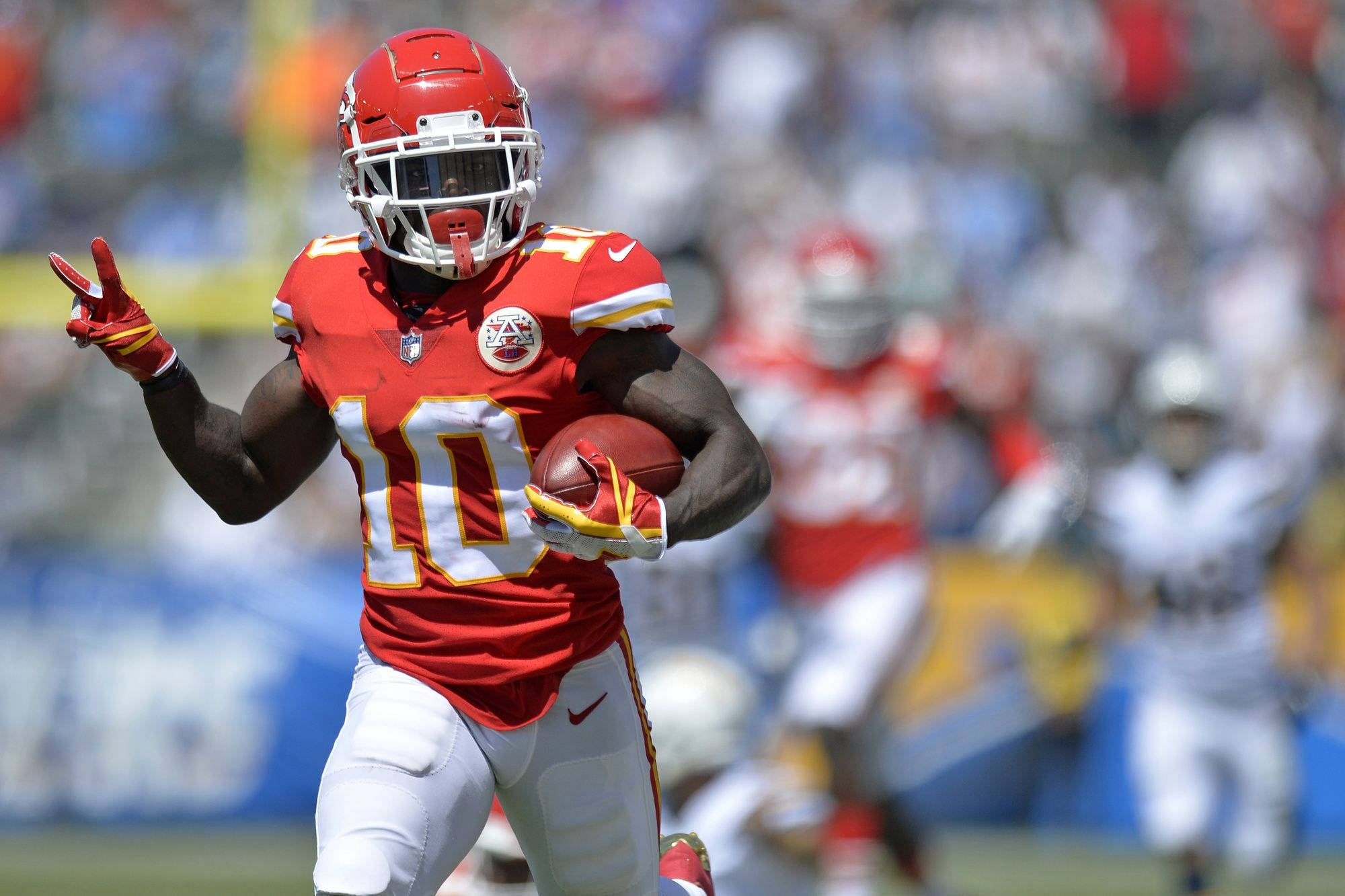 Tyreek Hill 桌布 壁紙 — 一名身穿紅色10號球衣的堪薩斯城酋長隊美式足球員在陽光明媚的比賽中帶球奔跑並做出和平手勢。