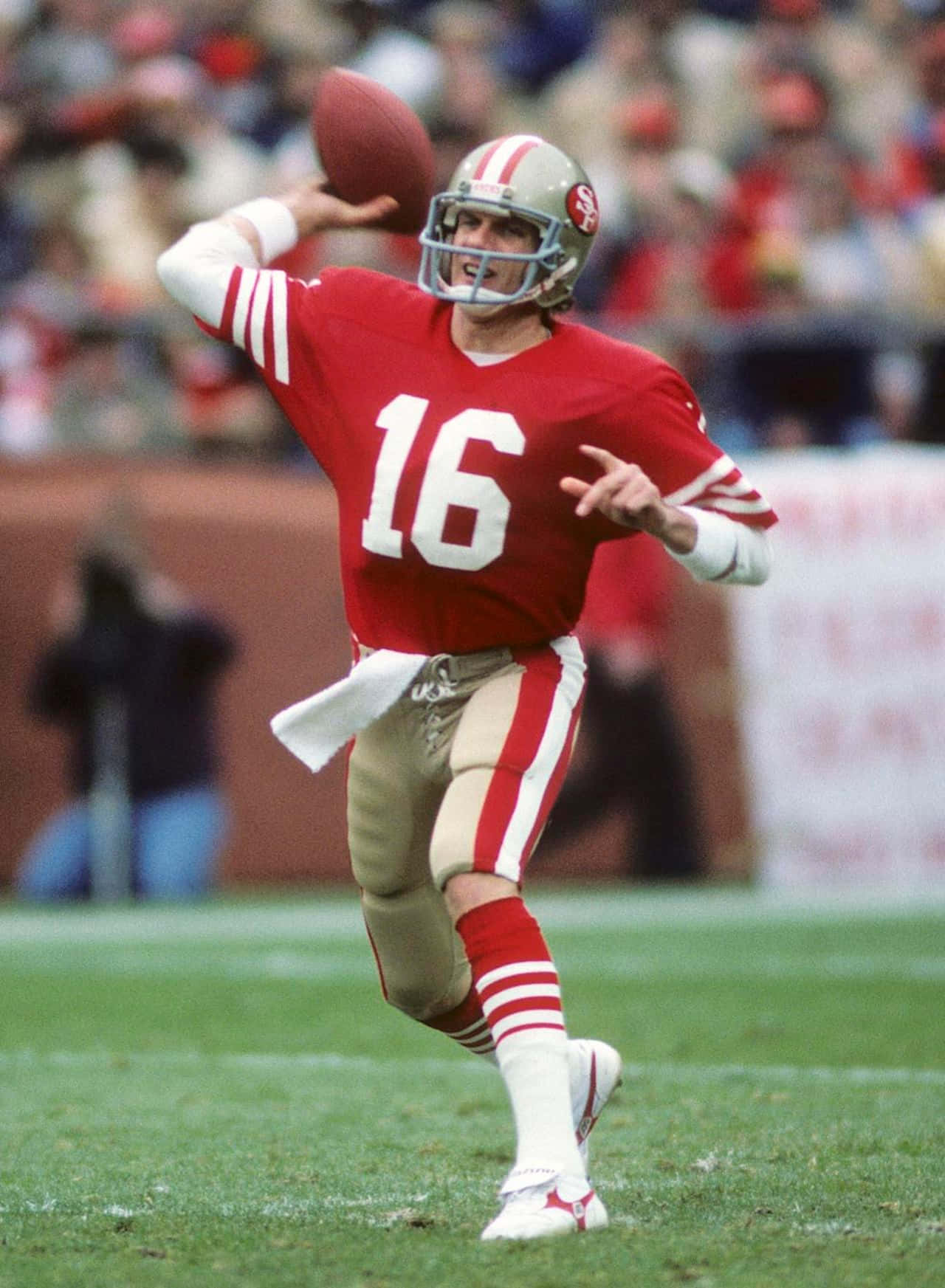 Joe Montana 桌布 壁紙 — 一名身穿紅色16號球衣的四分衛在日間比賽中擲出橄欖球，背景是模糊的人群。