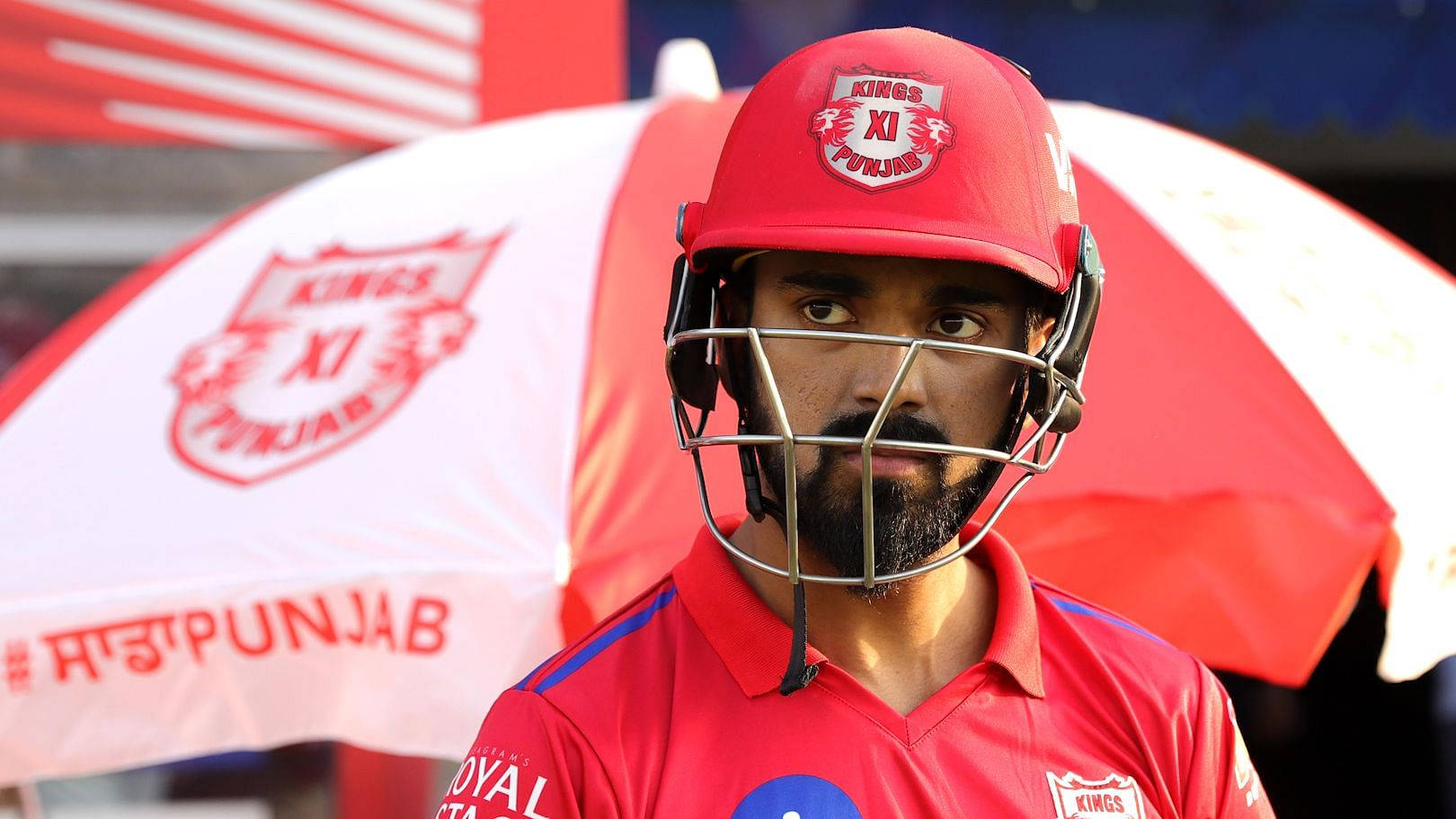 punjab kings 桌布 壁紙 — 一名身穿紅色Kings XI Punjab裝備的板球運動員，在品牌紅白傘下神情專注地站立。