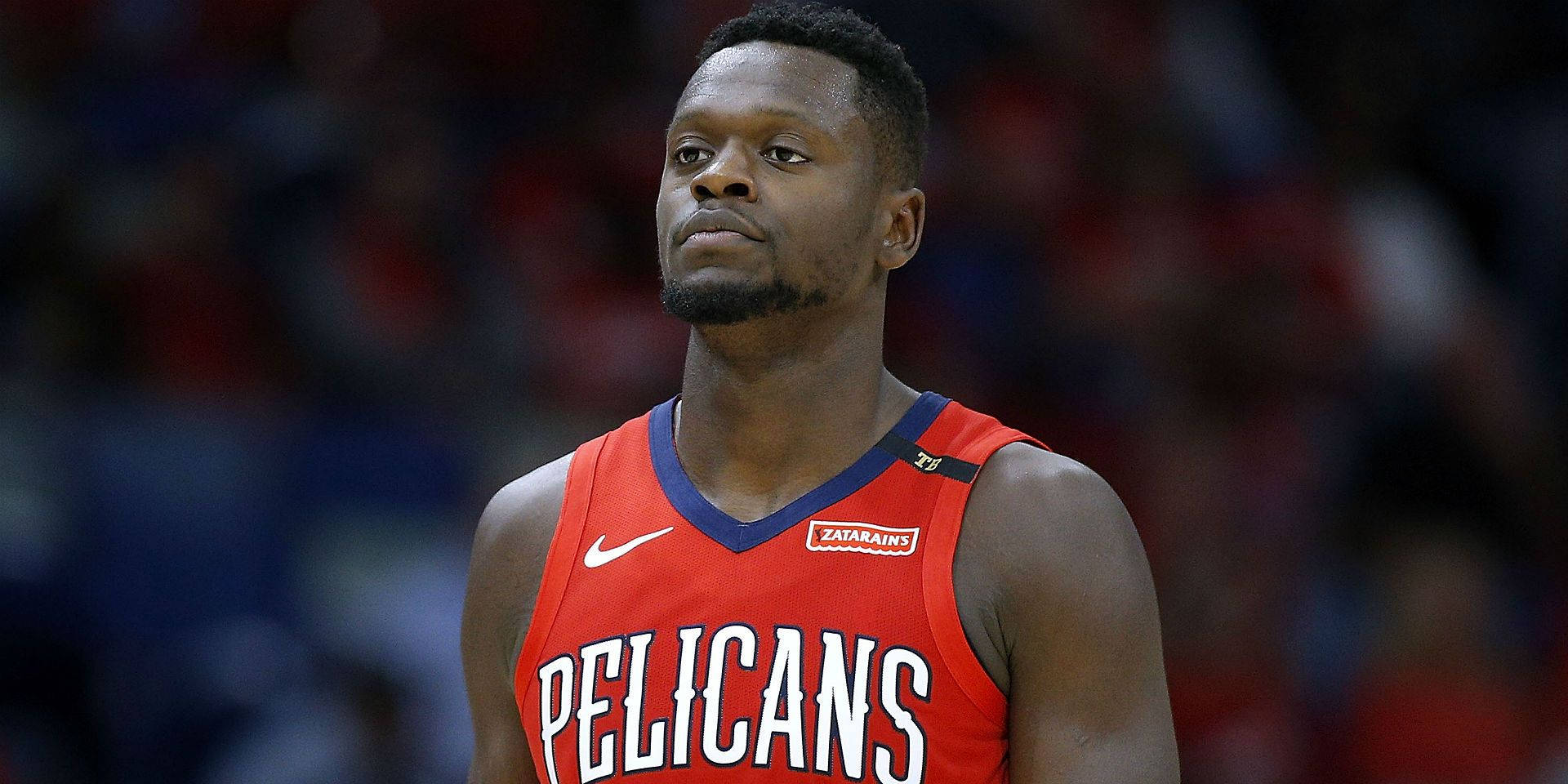 運動 桌布 壁紙 — 一名身穿紅色Pelicans球衣、專注的籃球運動員站在球場上，背景是模糊的觀眾。