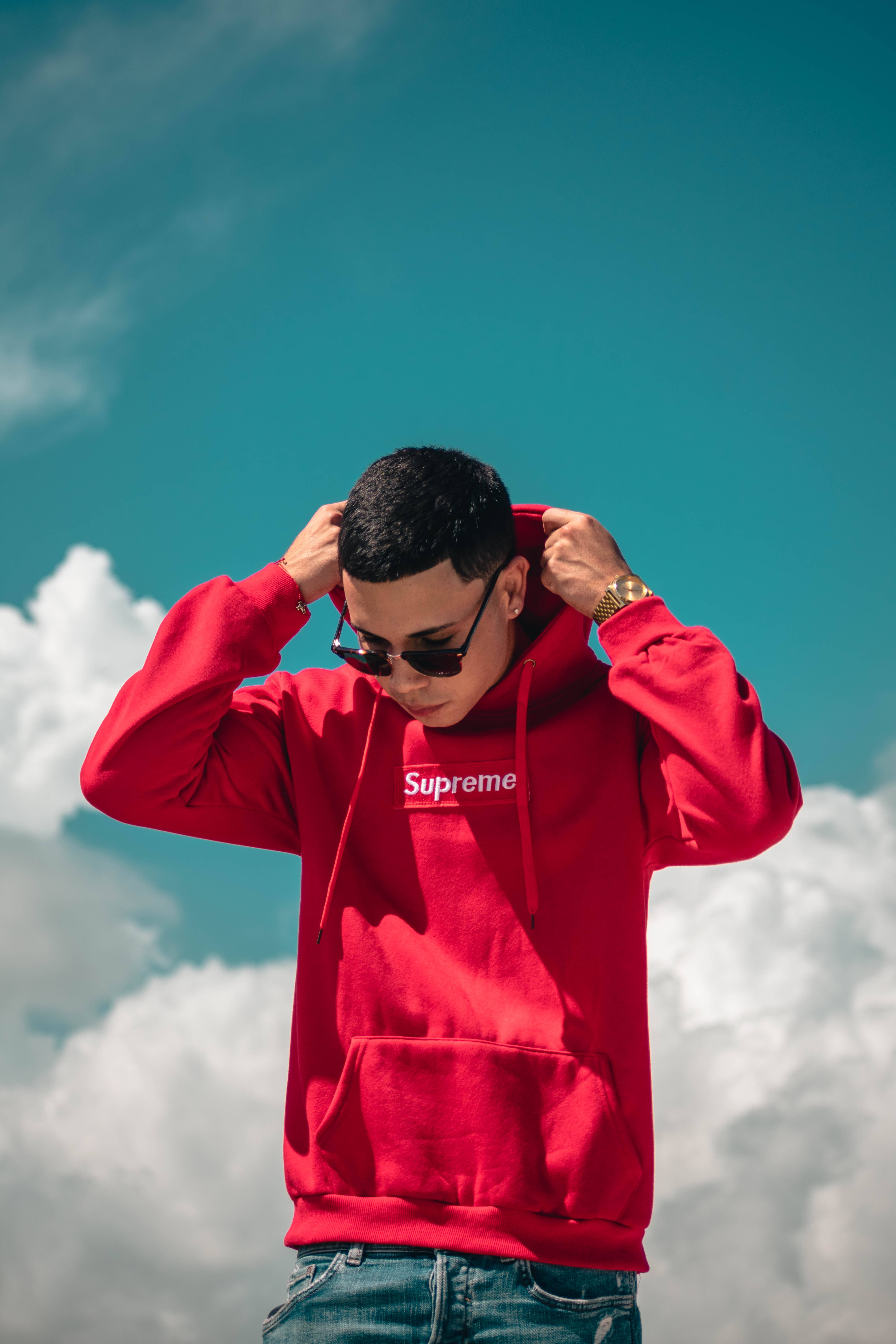 supreme 桌布 壁紙 — 一名身穿紅色Supreme連帽衛衣的年輕男子在藍天白雲下調整他的帽子。