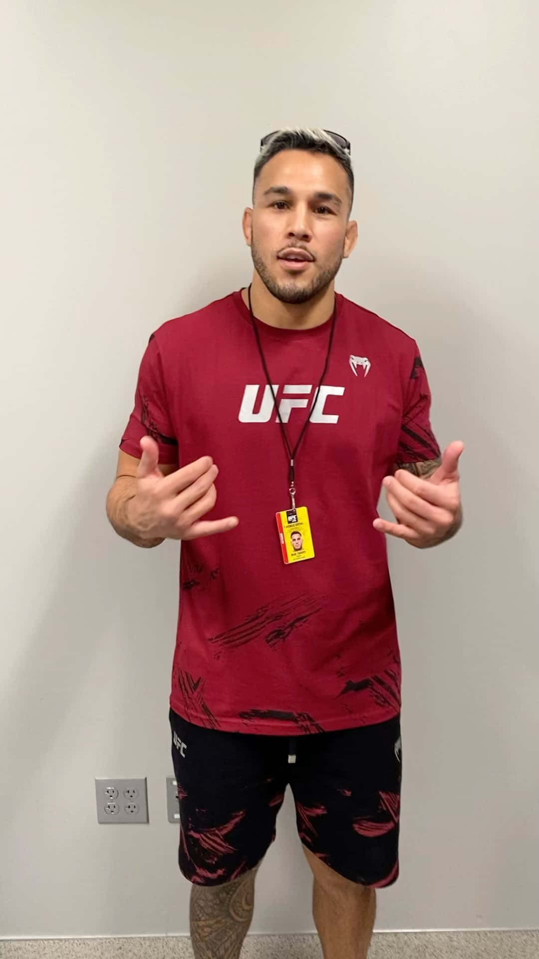 UFC 桌布 壁紙 — 一名身穿紅色UFC襯衫和黑色短褲的男子，脖子上掛著掛繩，站在純白色牆壁前做手勢。