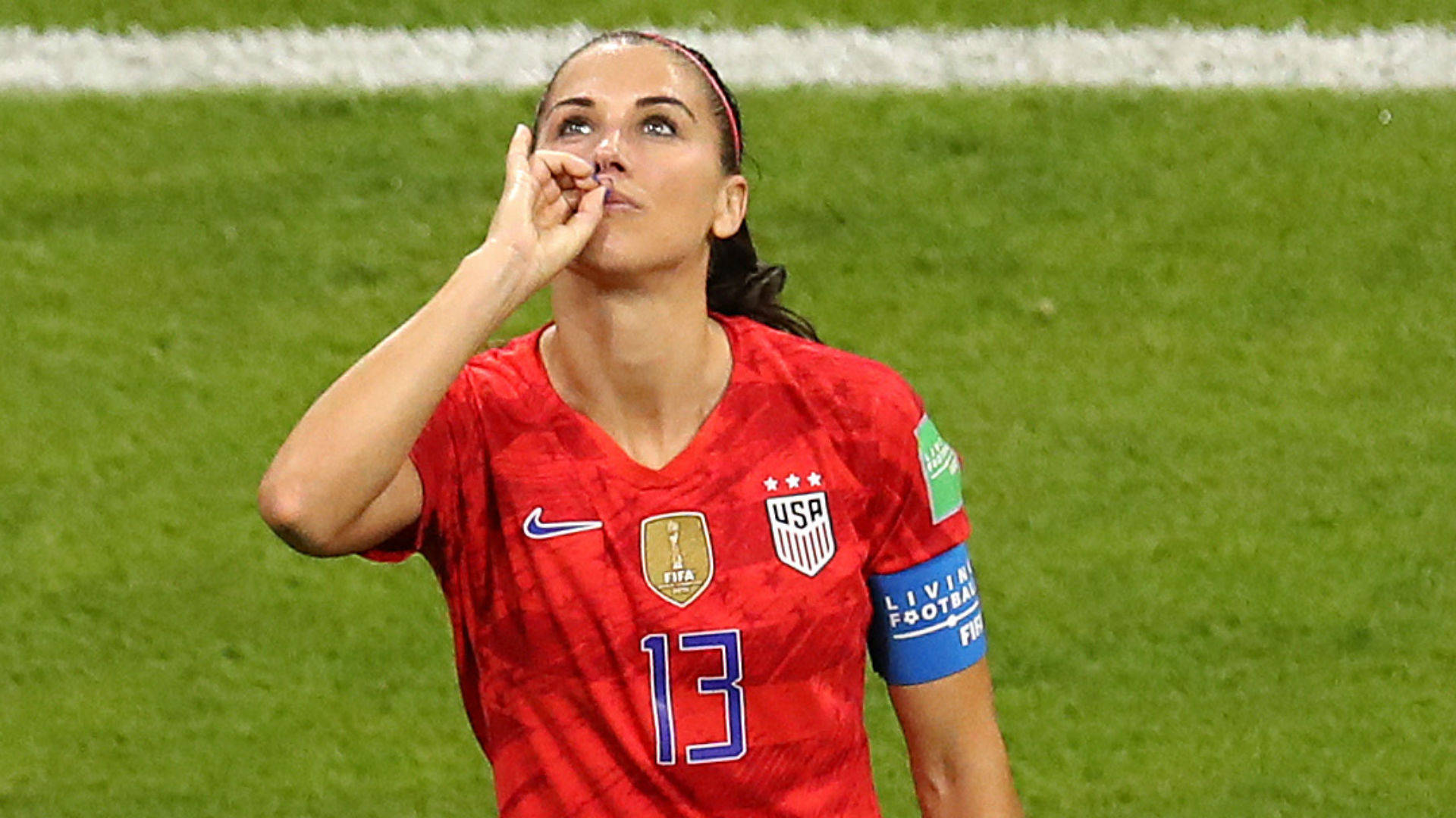 Alex Morgan 桌布 壁紙 — 一名身穿紅色USWNT球衣、號碼13的女足球員在綠色球場上，手靠近臉部向上看。