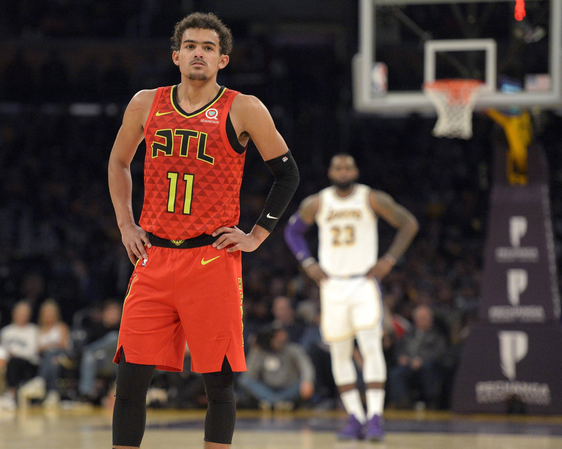 Trae Young 桌布 壁紙 — 一名身穿紅色亞特蘭大球衣的籃球運動員在場館燈光下的球場上雙手叉腰站立。