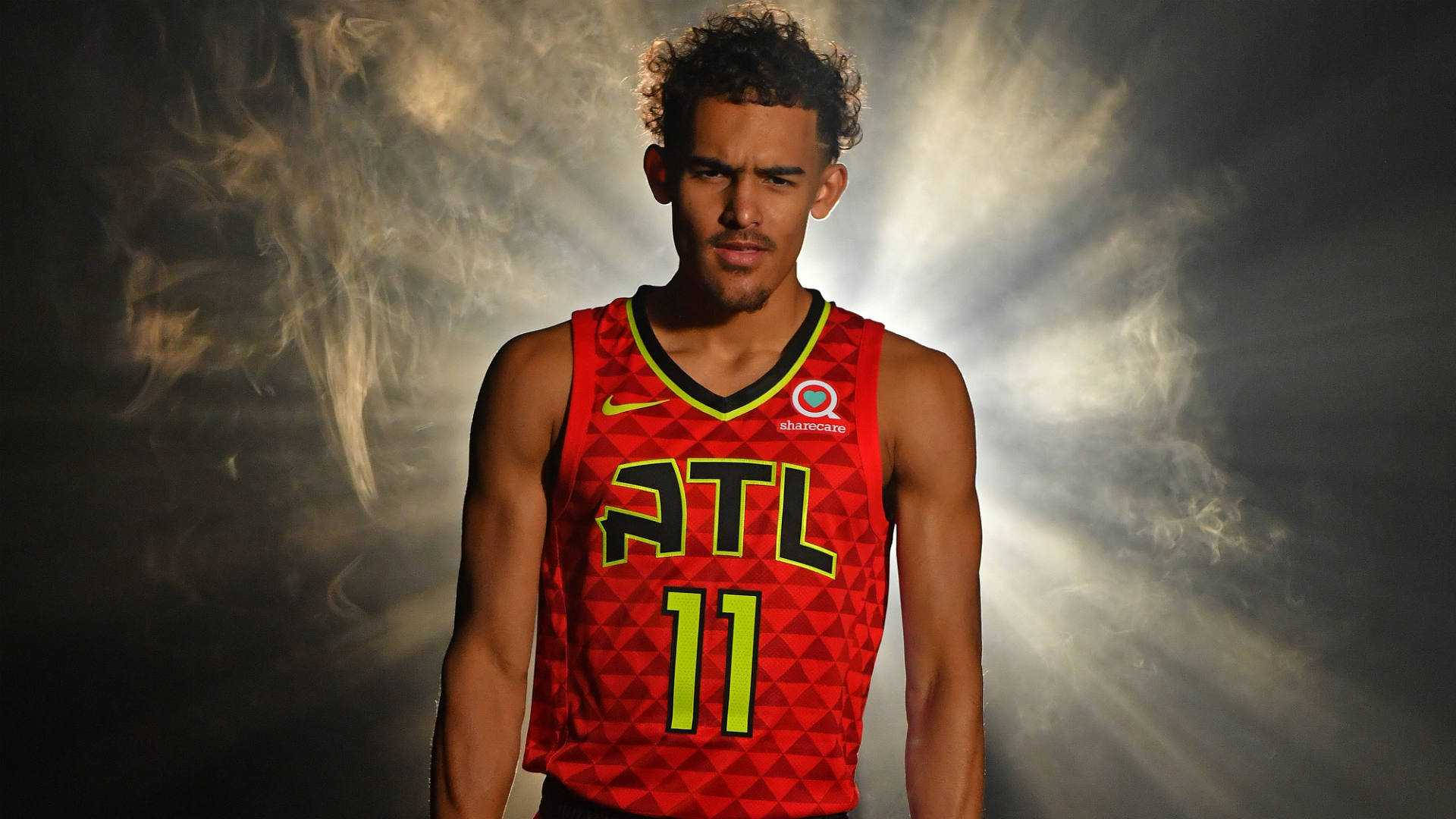 Trae Young 桌布 壁紙 — 一名身穿紅色亞特蘭大老鷹隊球衣的籃球運動員，自信地站在煙霧瀰漫、光線四射的戲劇性背景前。