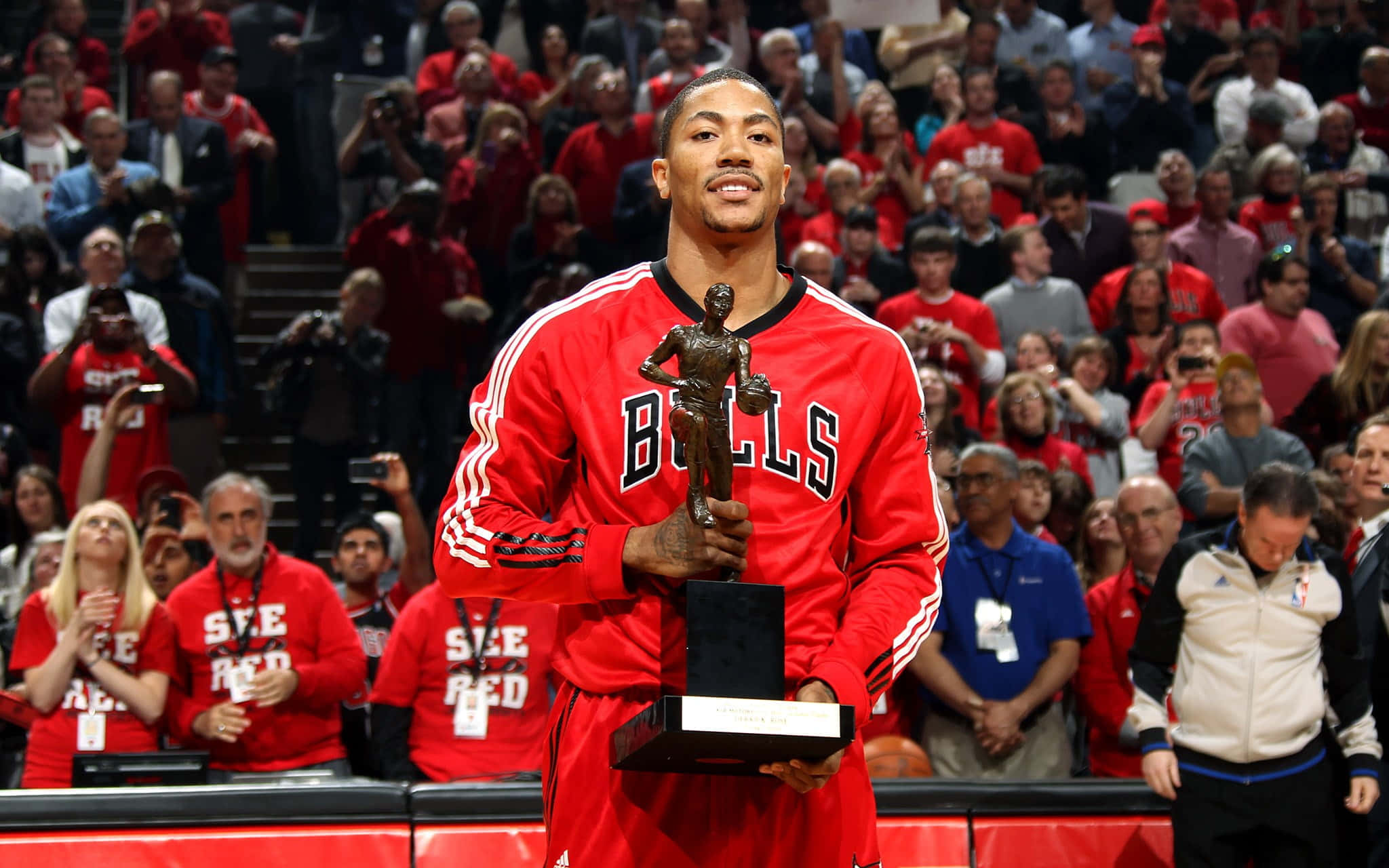derrick rose 桌布 壁紙 — 一名身穿紅色公牛隊球衣的籃球運動員在球場上微笑著高舉獎盃，背景是歡呼的球迷。
