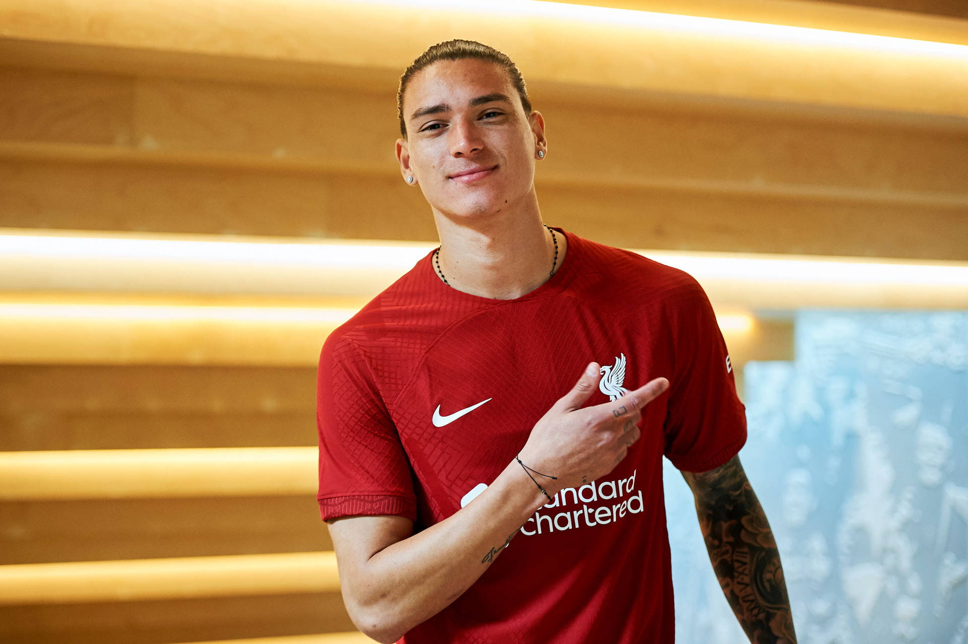 Nike 桌布 壁紙 — 一名身穿紅色利物浦FC球衣的年輕男子微笑著指向俱樂部徽章，背景是溫暖的木鑲板。