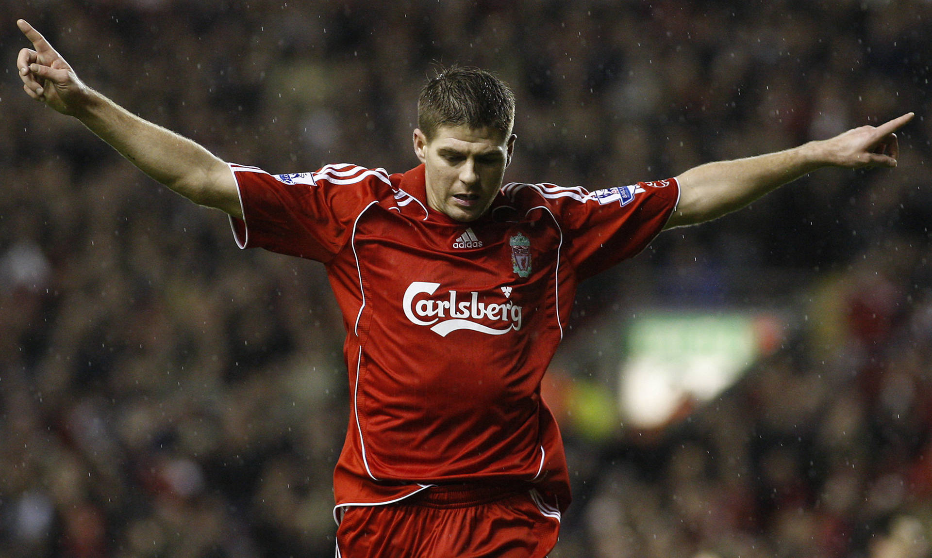 Steven Gerrard 桌布 壁紙 — 一名身穿紅色利物浦球衣的足球員在夜間比賽中於雨中張開雙臂慶祝。