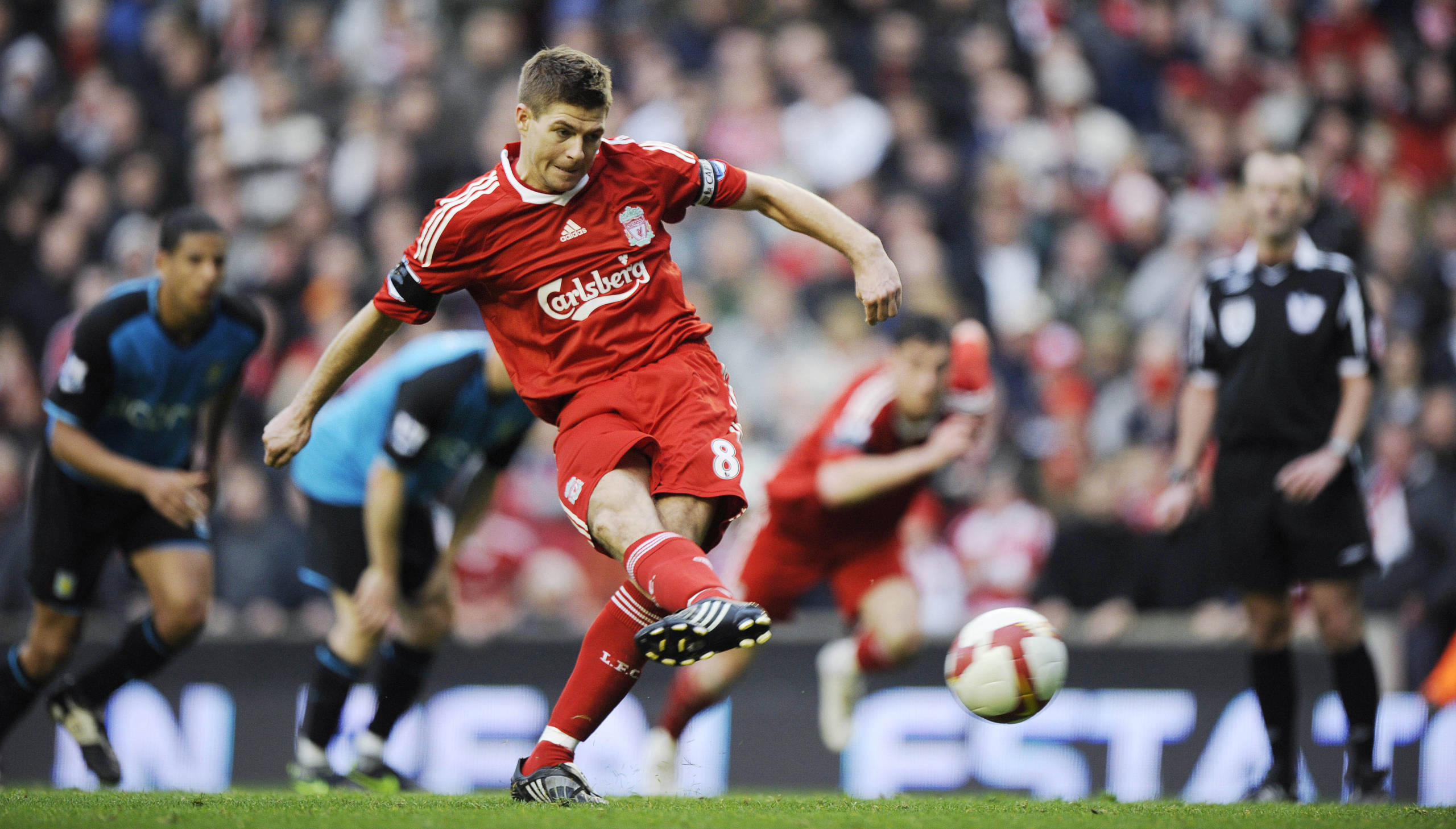 Steven Gerrard 桌布 壁紙 — 一名身穿紅色利物浦球衣的足球員在比賽中踢球，背景是模糊的隊友和人群。