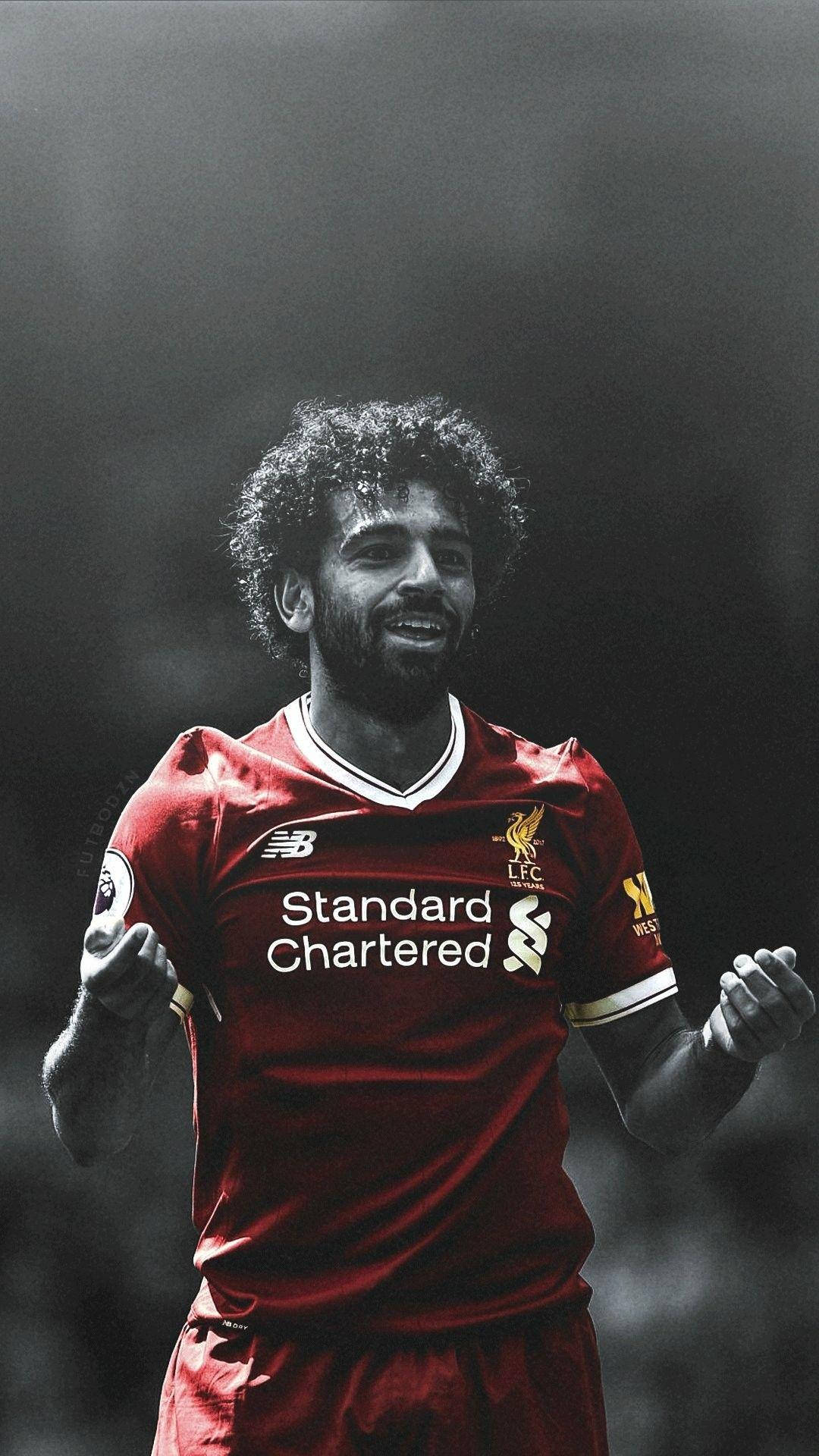 Salah 桌布 壁紙 — 一名身穿紅色利物浦球衣的足球運動員在黑暗模糊的球場背景下勝利地微笑。