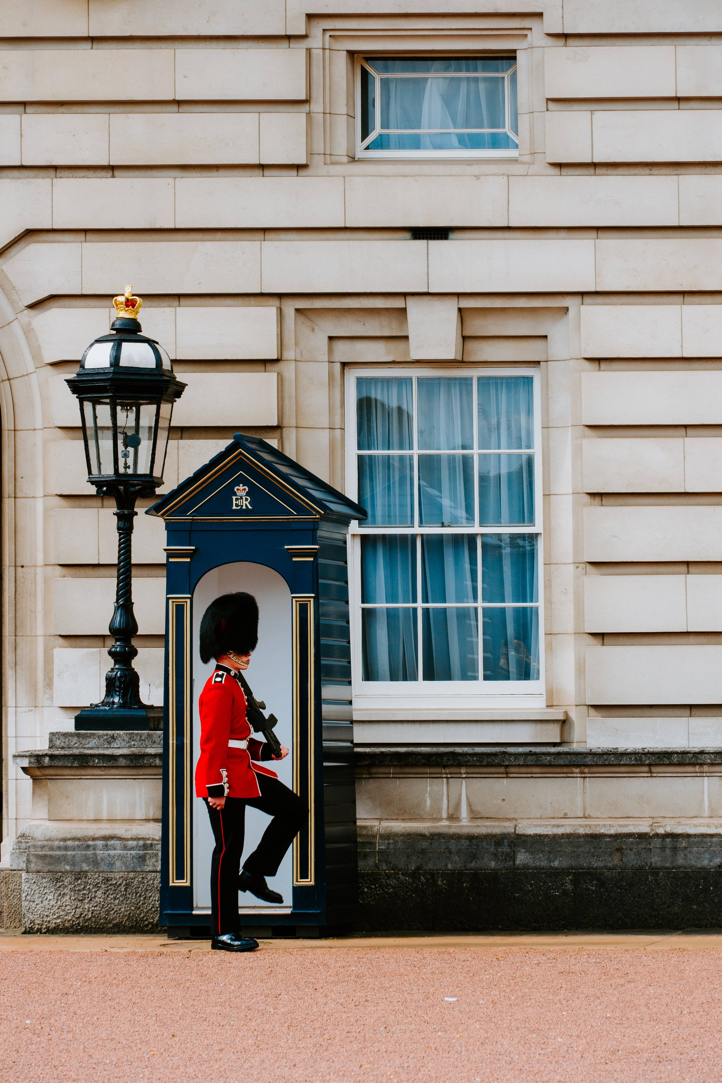 England 桌布 壁紙 — 一名身穿紅色制服的王室衛兵，在藍色崗亭旁立正，背景是米色石建築物帶有窗戶。