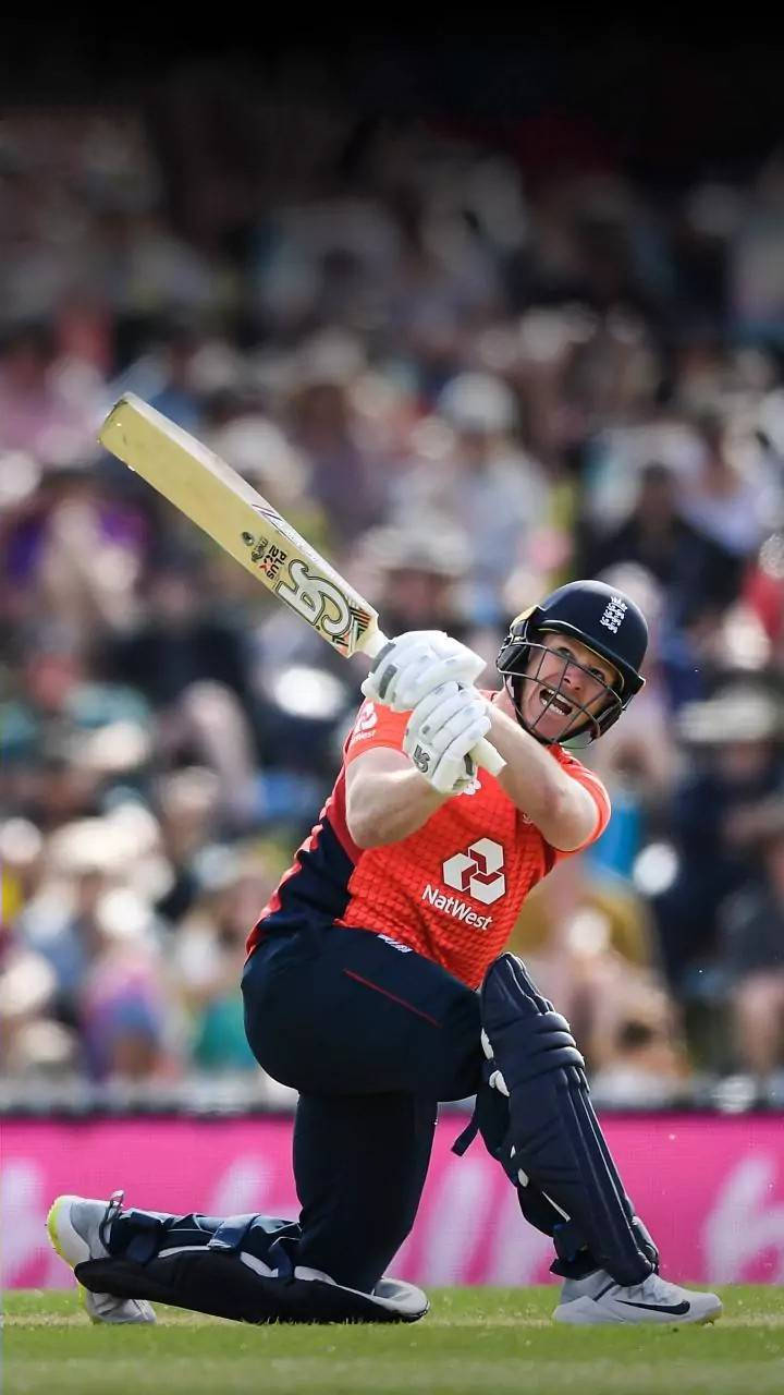 eoin morgan 桌布 壁紙 — 一名身穿紅色和深藍色裝備的板球擊球手在日間比賽中強力揮棒，背景是模糊的人群。